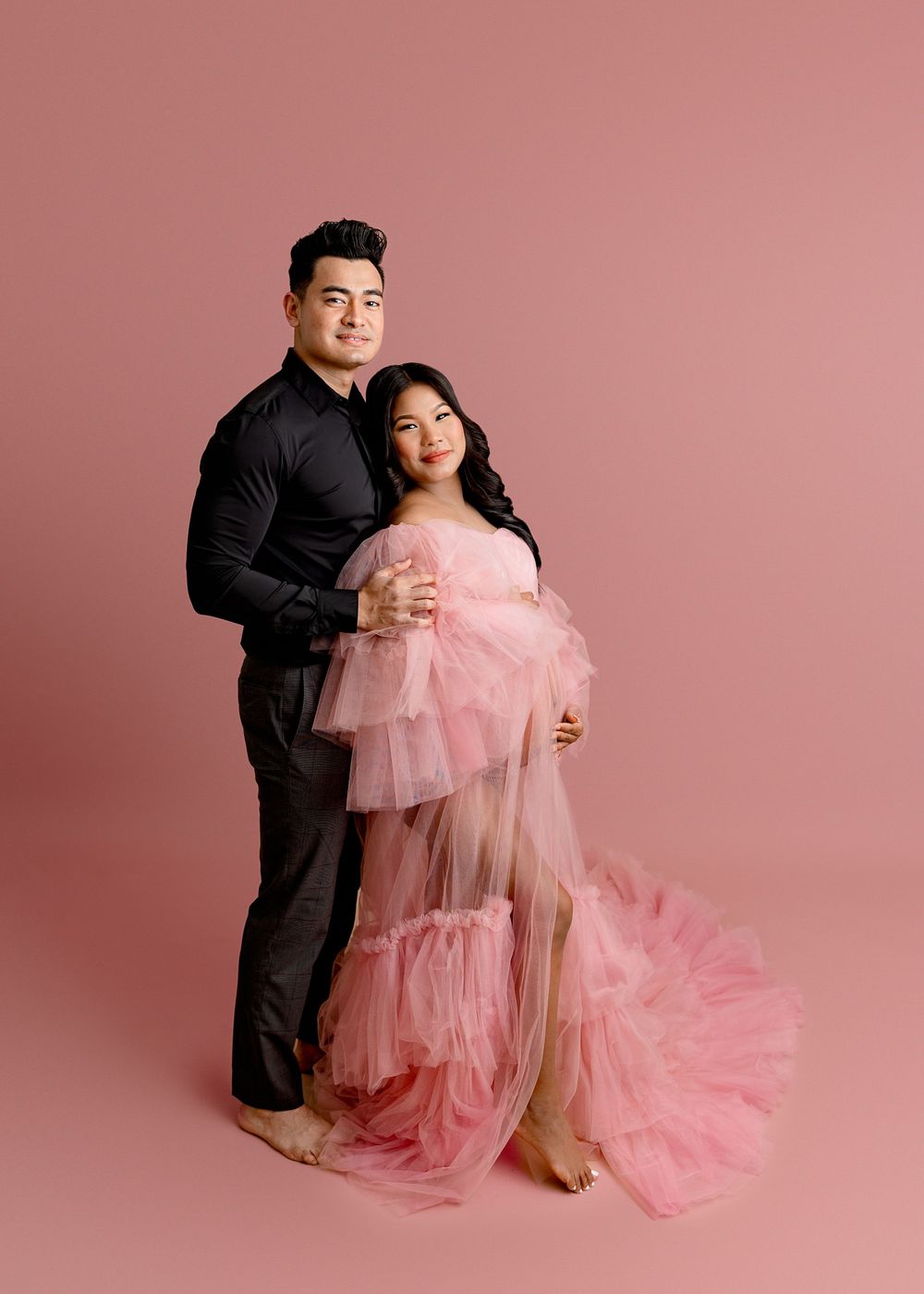 Romantic maternity portrait with couple in pink tulle gown against rose backdrop.