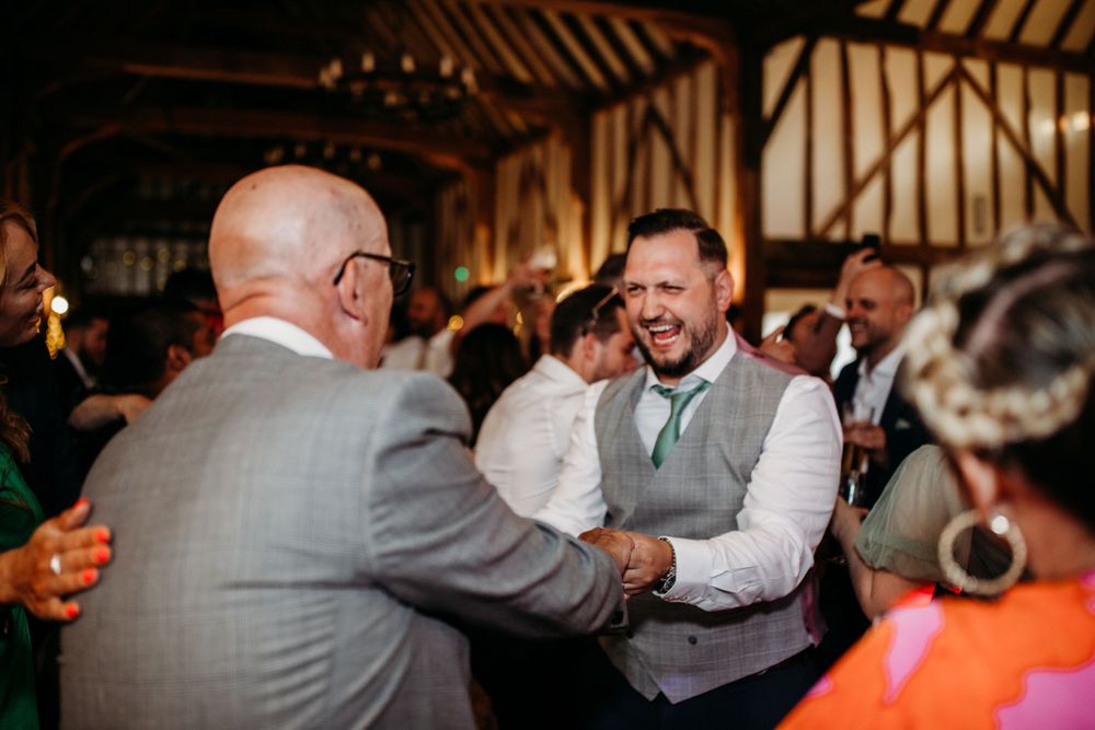 Two guests share an energetic handshake and laugh during wedding reception celebrations.