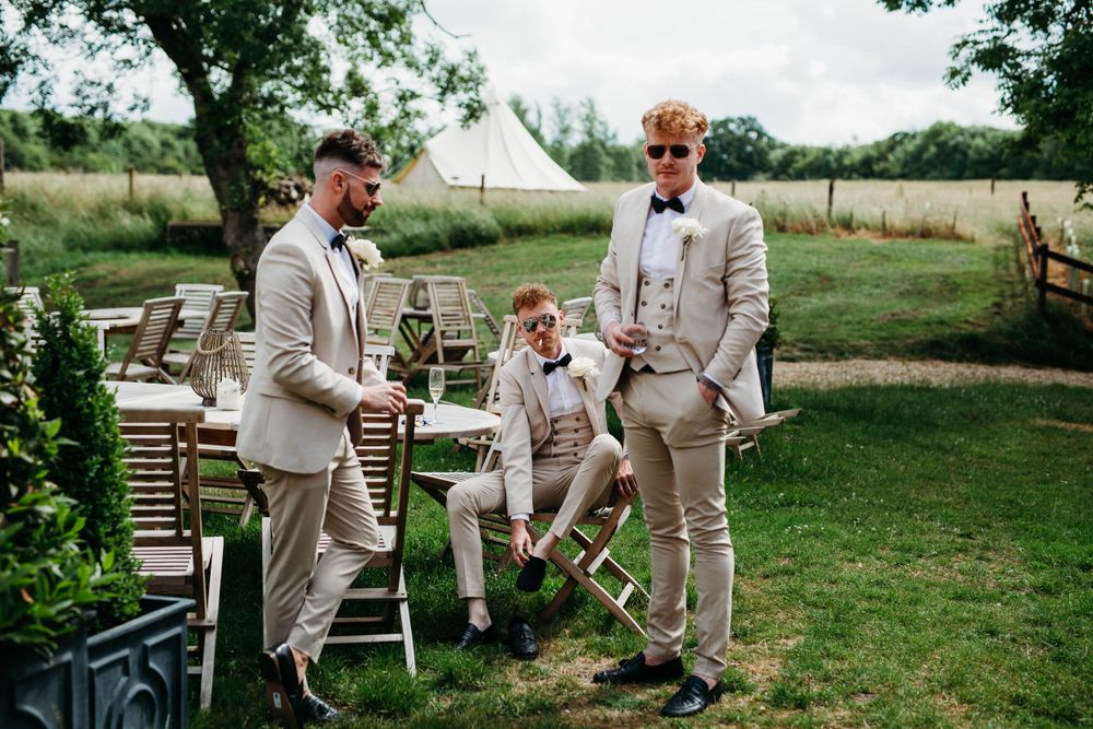 Wedding party in beige suits laughs together on lawn with white tent in background.