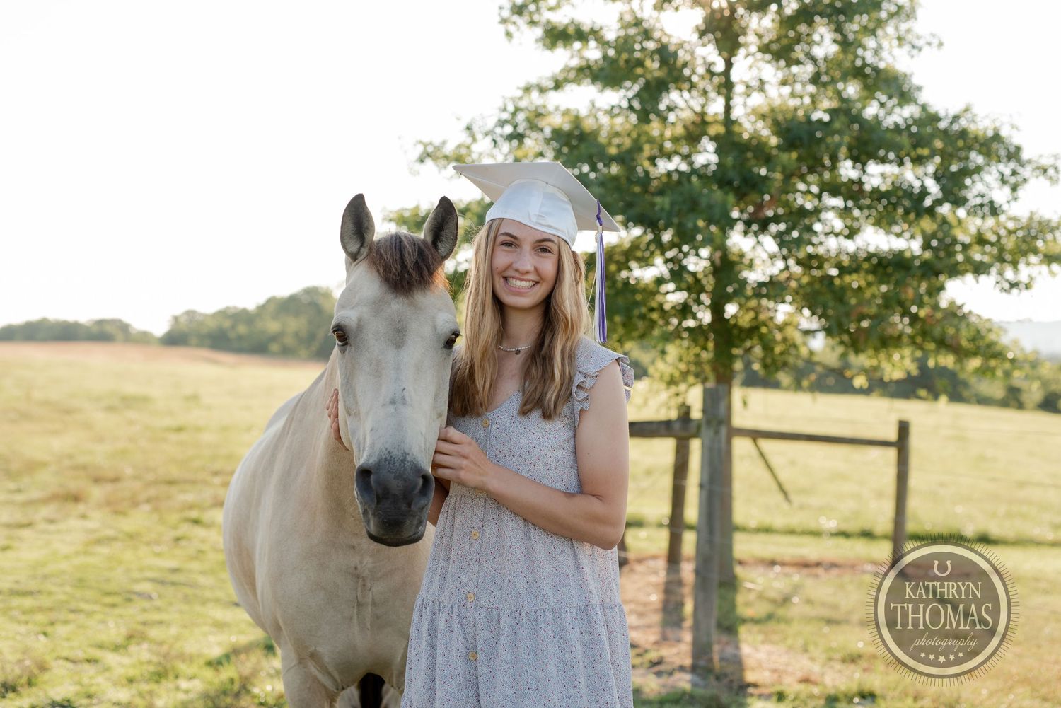 Graduate in a light dress poses with a white horse in a pastoral field setting.
