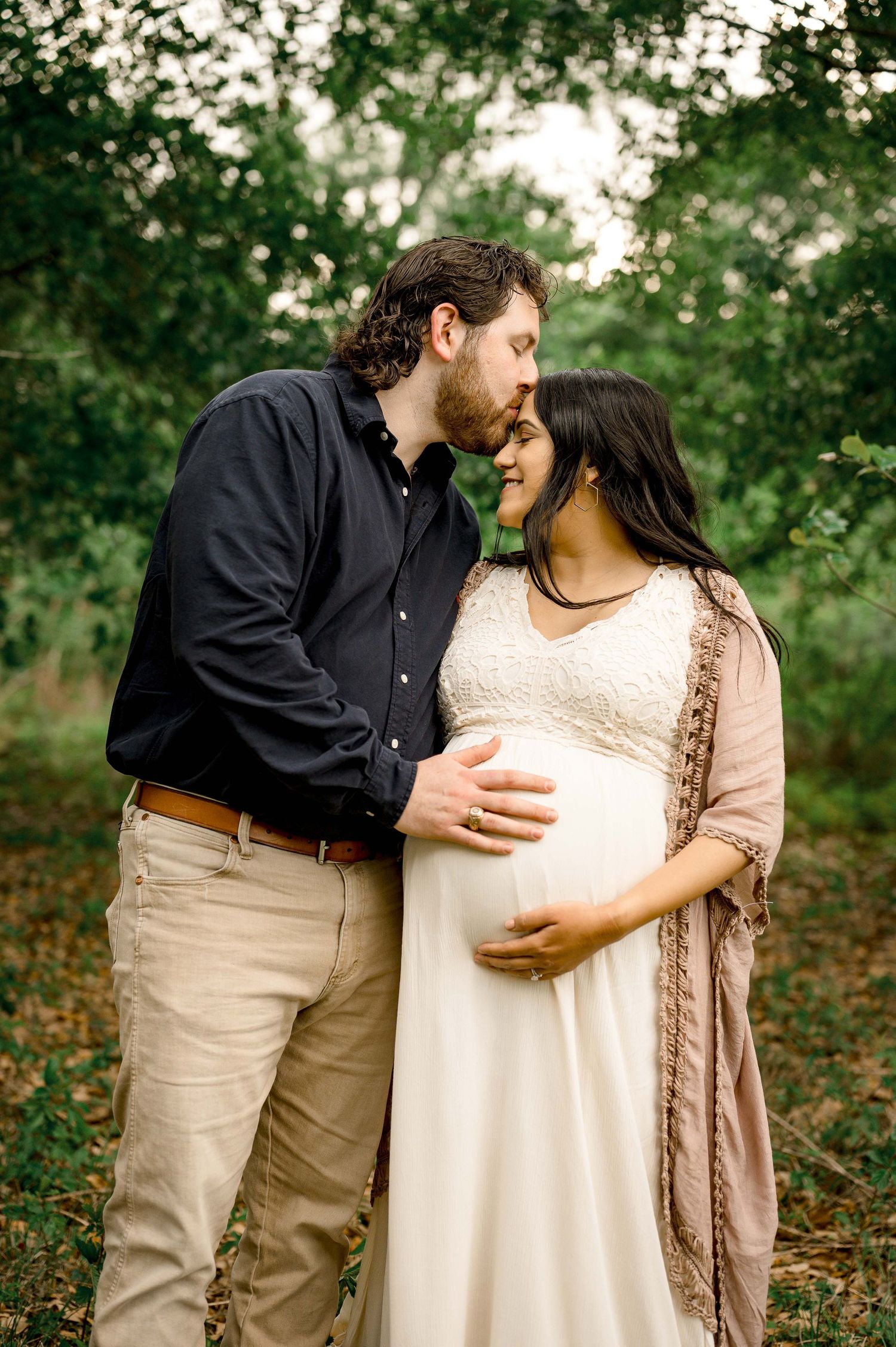husband kissing his wife forehead while touching his wife belly during a maternity session in the woods