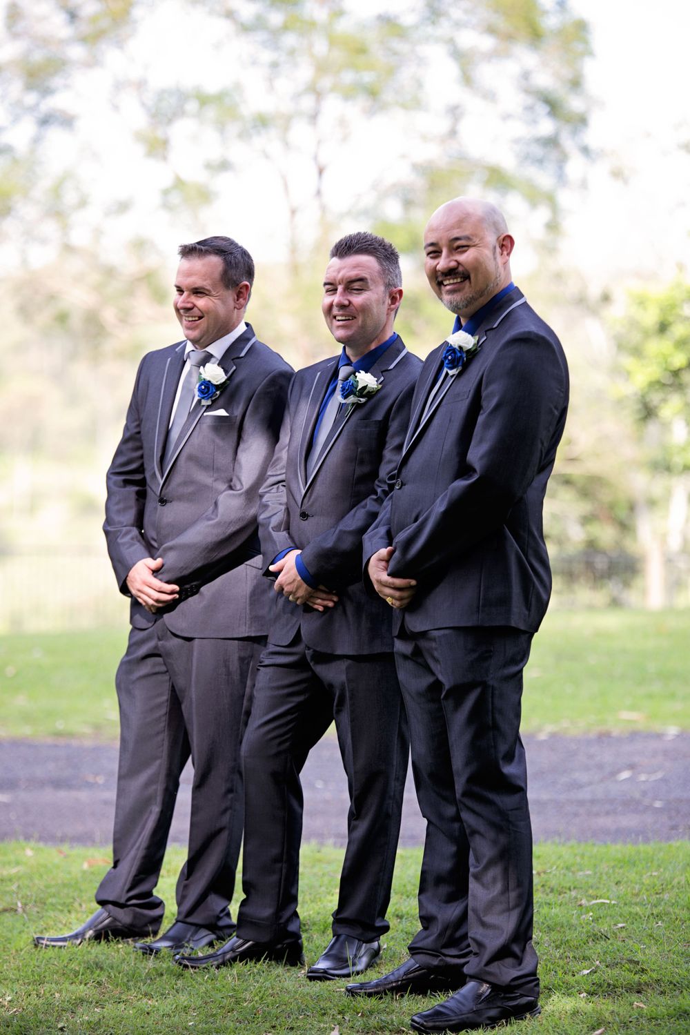 Groomsmen in dark suits with blue accents pose together on grassy lawn.