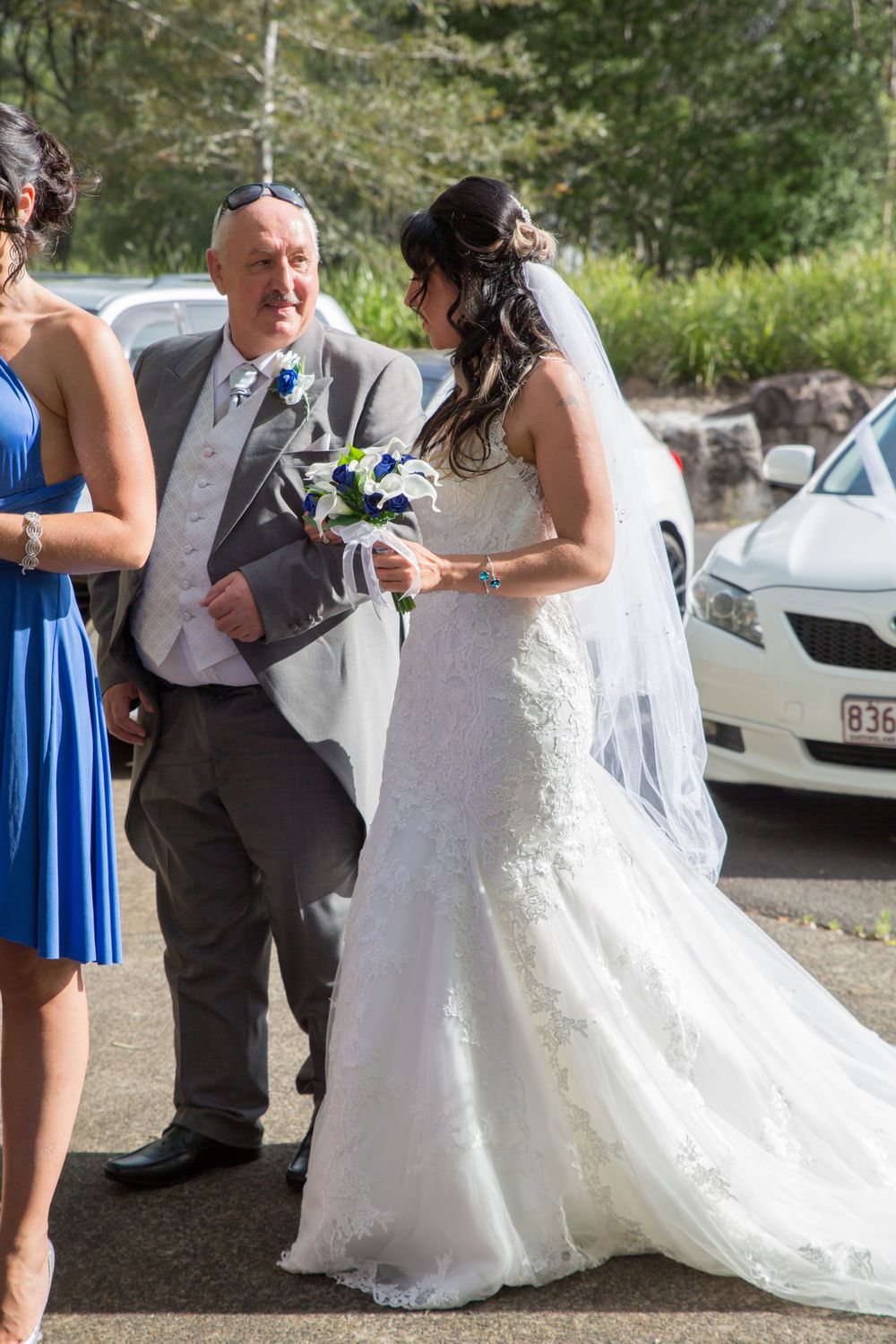 A wedding party gathers outside with the bride wearing a flowing white lace gown and veil while holding a bouquet.