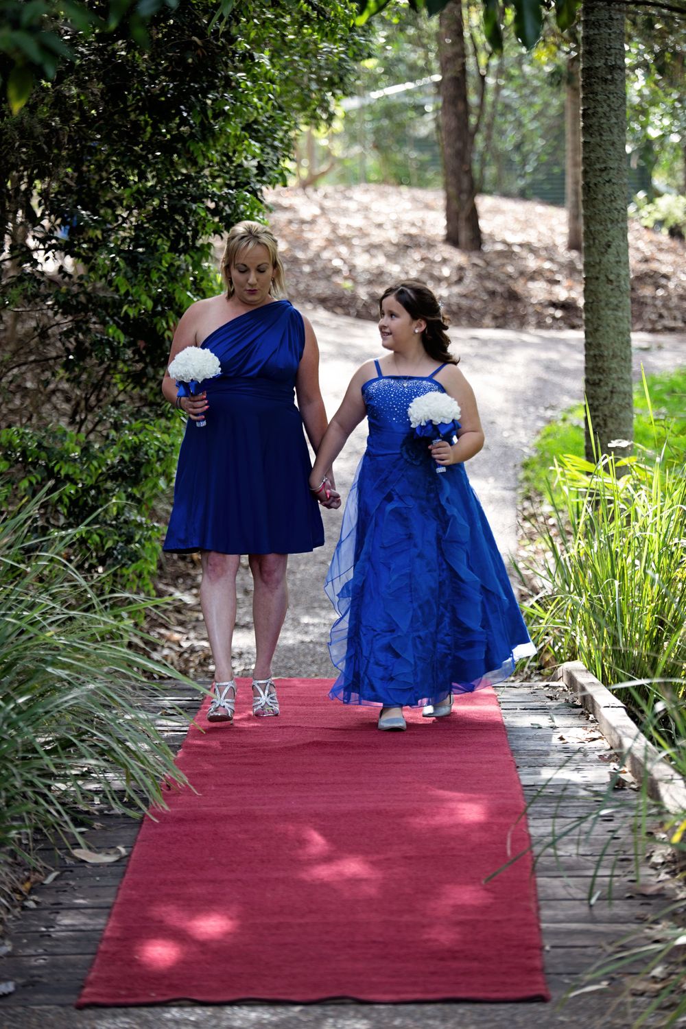 Two bridesmaids in royal blue dresses walk down a red carpeted garden path holding white bouquets.