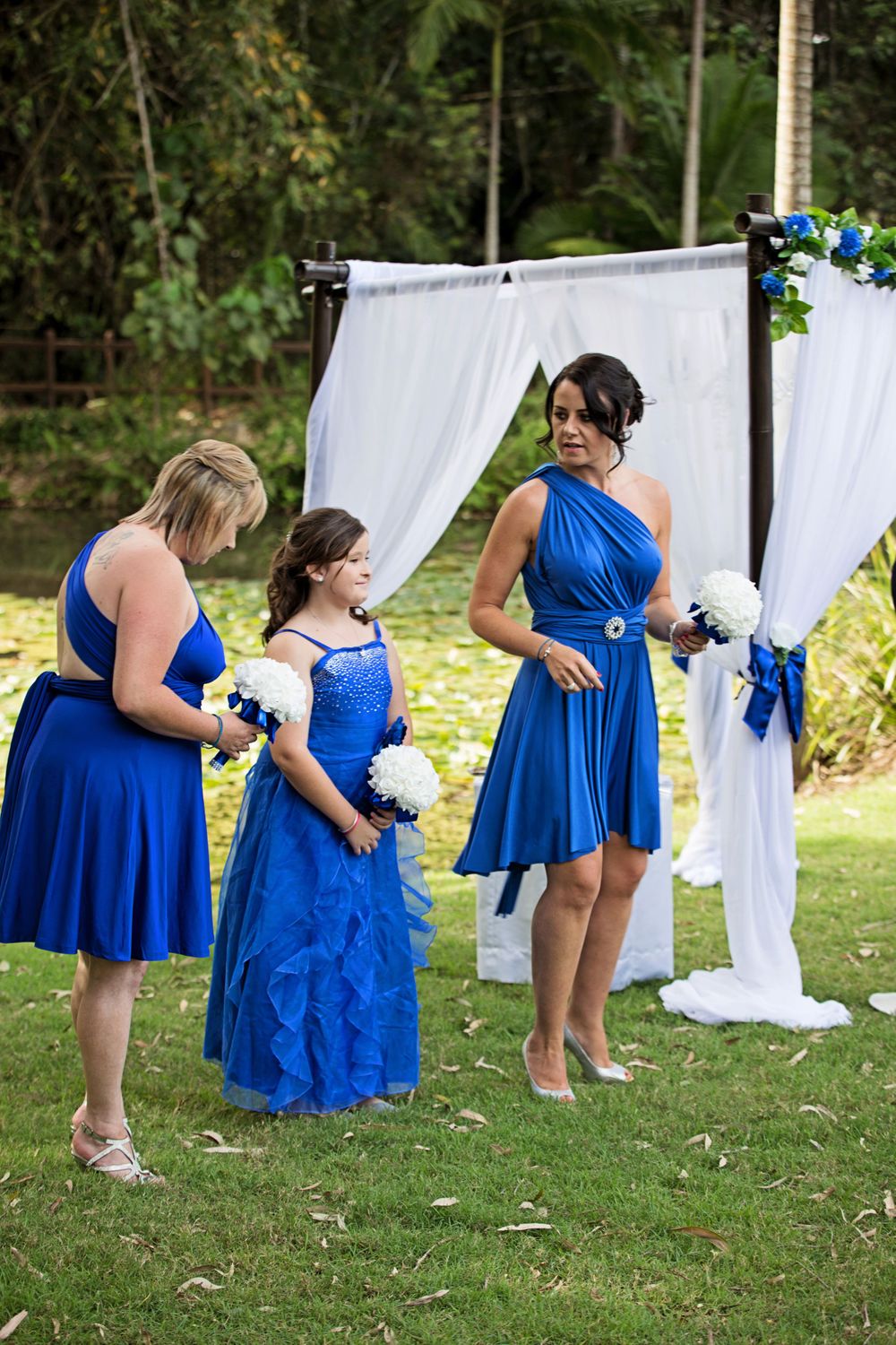 Three women in coordinating royal blue dresses stand together at an outdoor wedding ceremony.