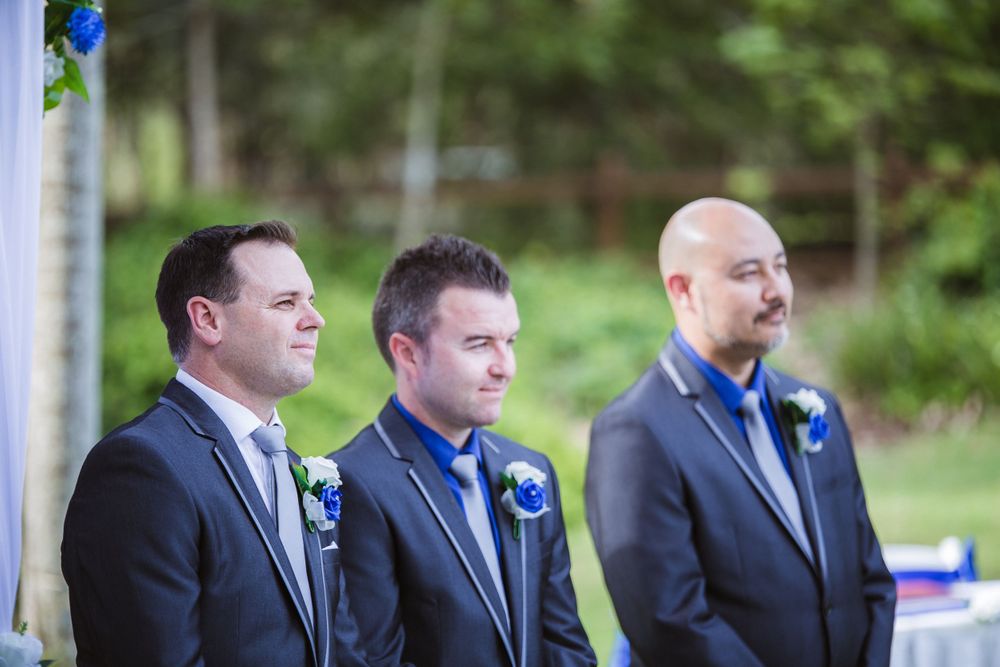 Three groomsmen in dark suits with blue accents stand at attention during an outdoor ceremony.