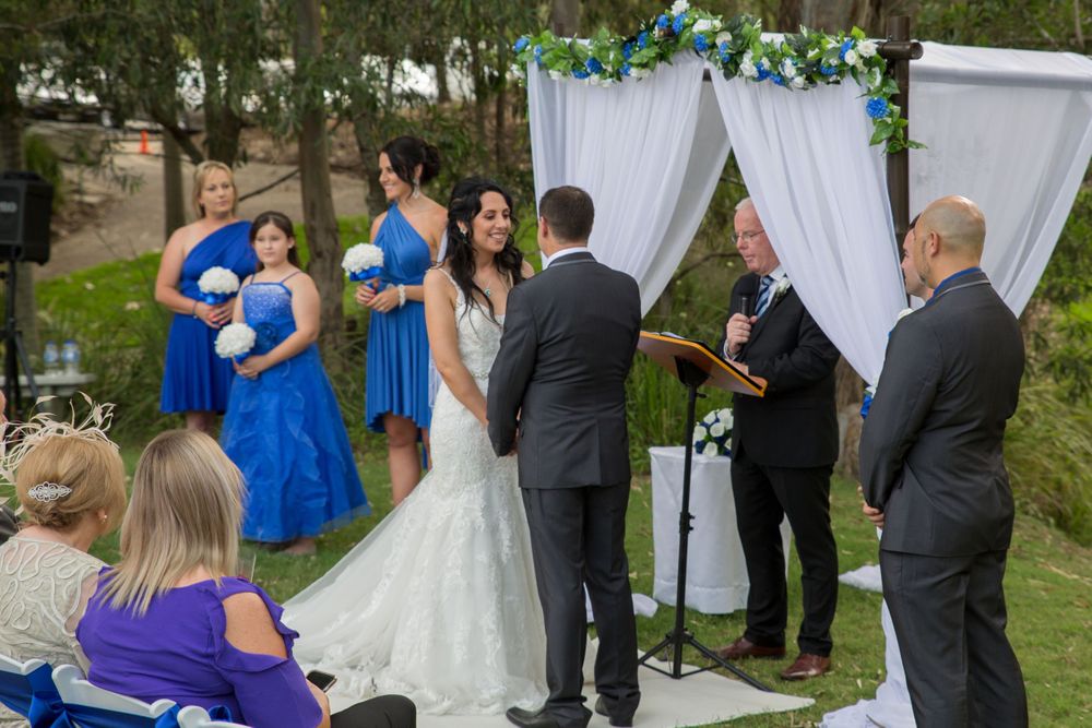 An outdoor wedding ceremony takes place under white draping decorated with blue and green florals.