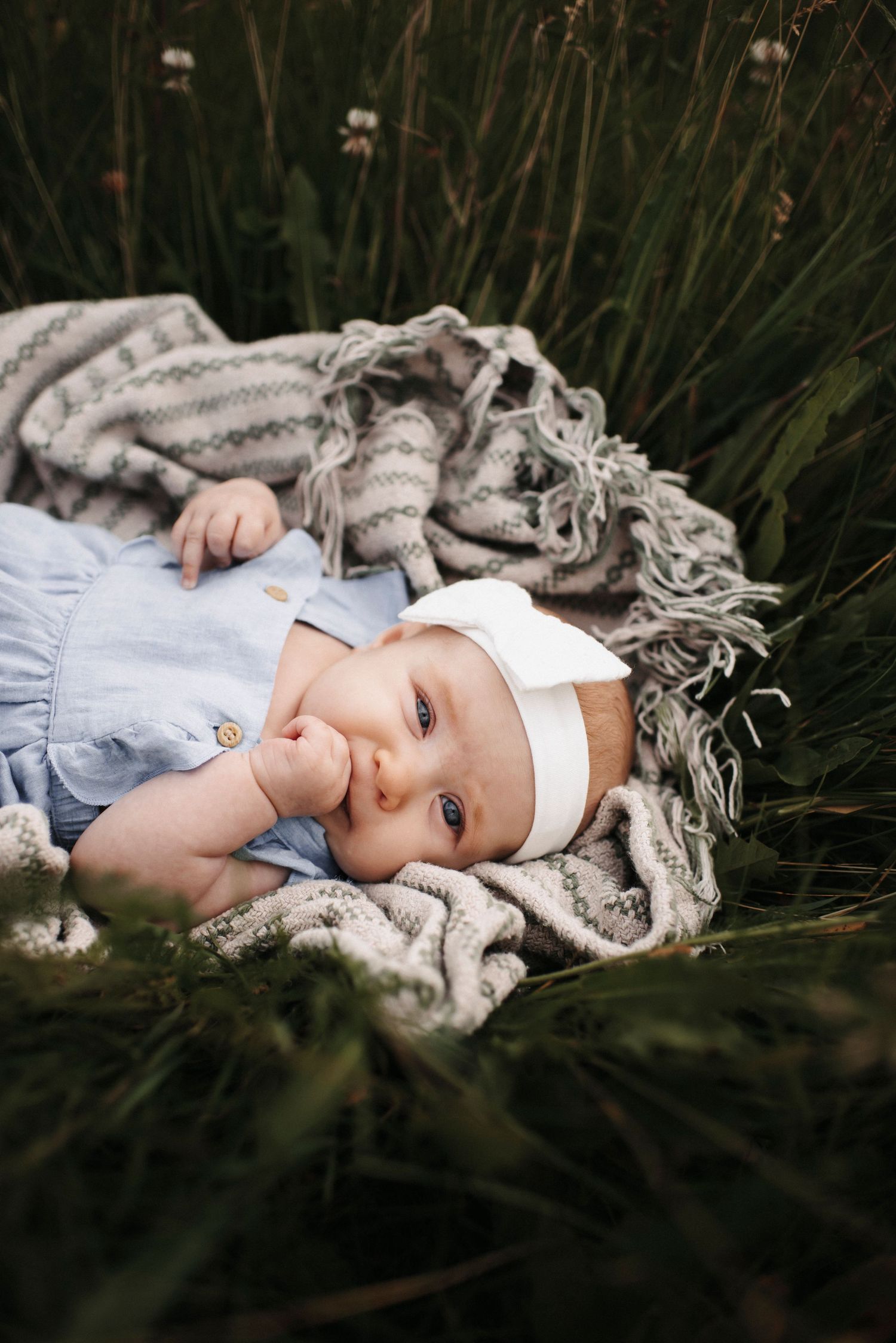Baby wearing white headband and blue outfit lies on striped blanket among white flowers.
