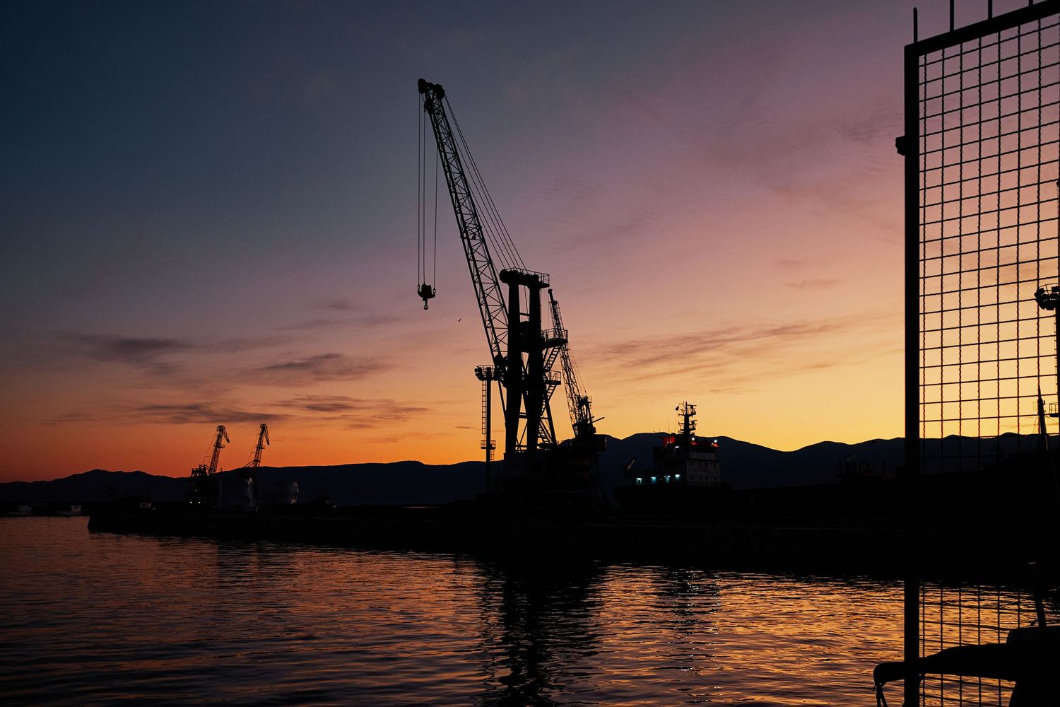 Industrial cargo cranes silhouetted against an orange sunset at a harbor.