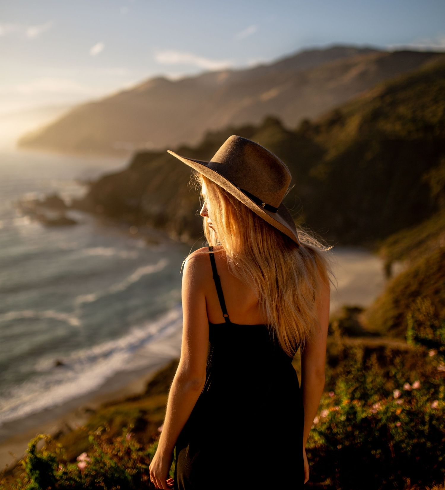 Person in dark dress and hat overlooking a scenic coastal landscape at sunset.