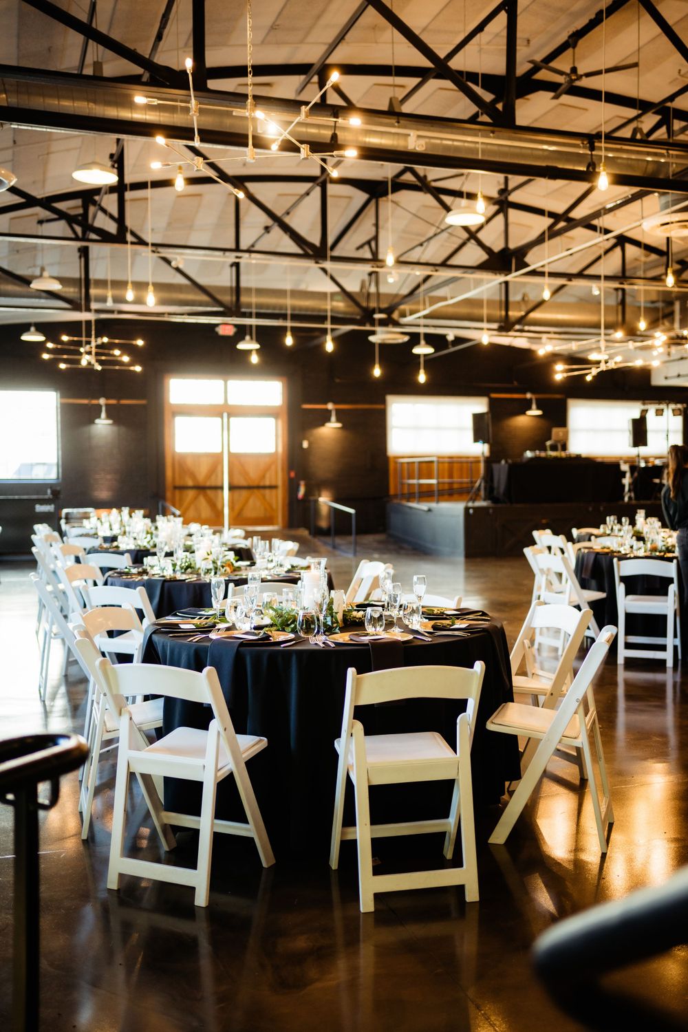 Modern reception venue with black tablecloths, white chairs, and industrial ceiling lights.