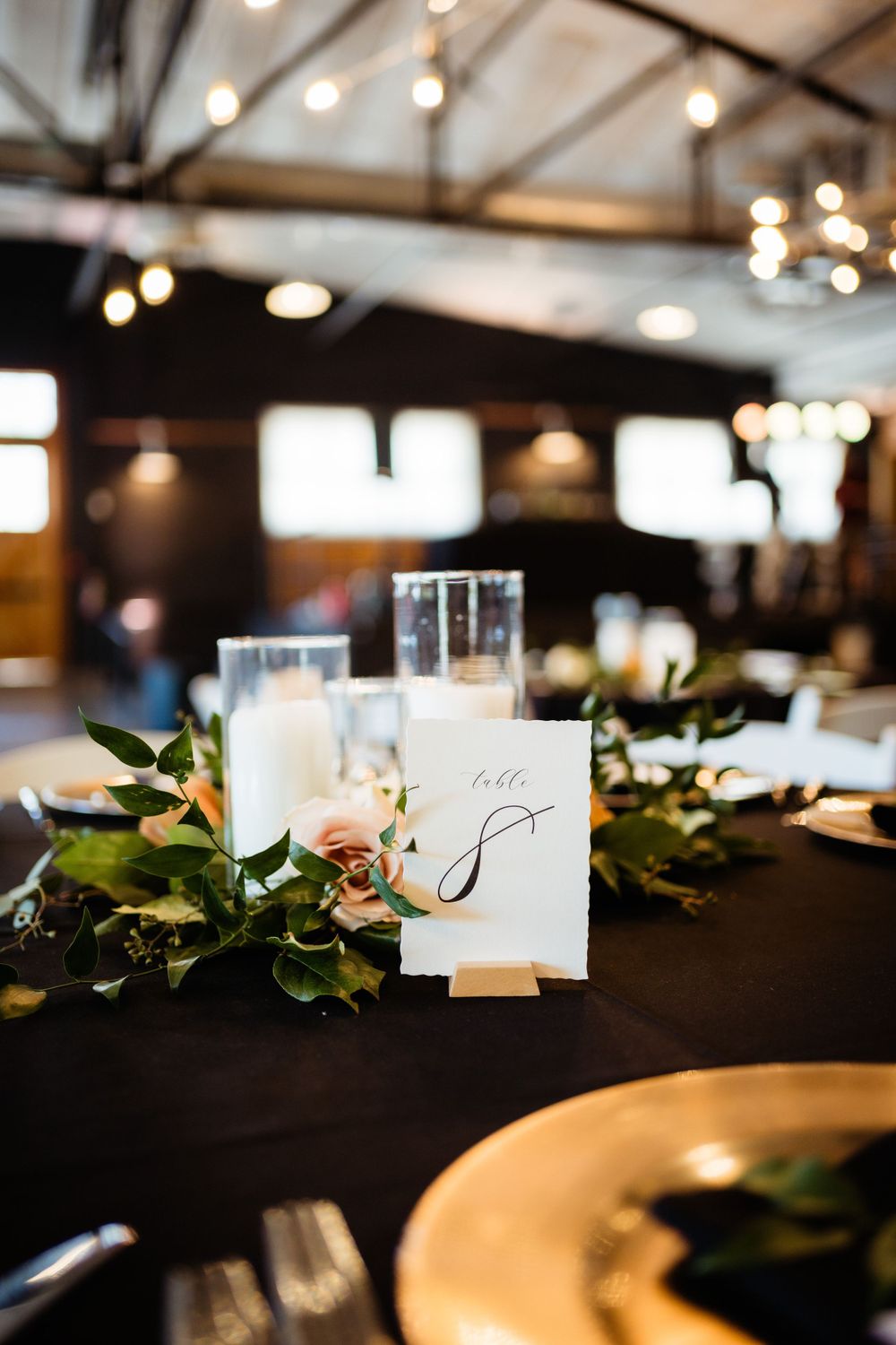 Elegant table setting with calligraphy table number and greenery on dark tablecloth.