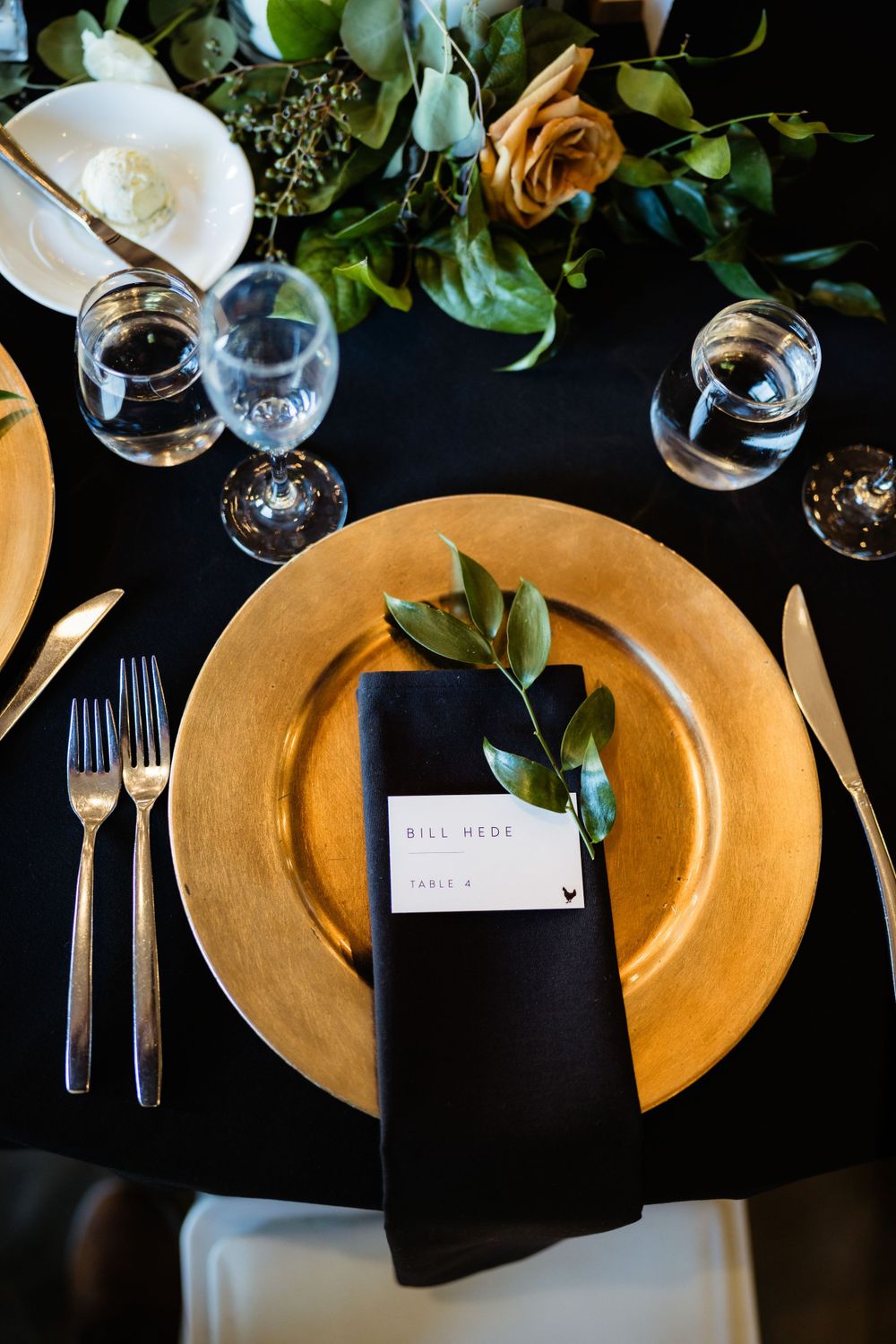 Elegant place setting with gold charger plate and black napkin on dark tablecloth.