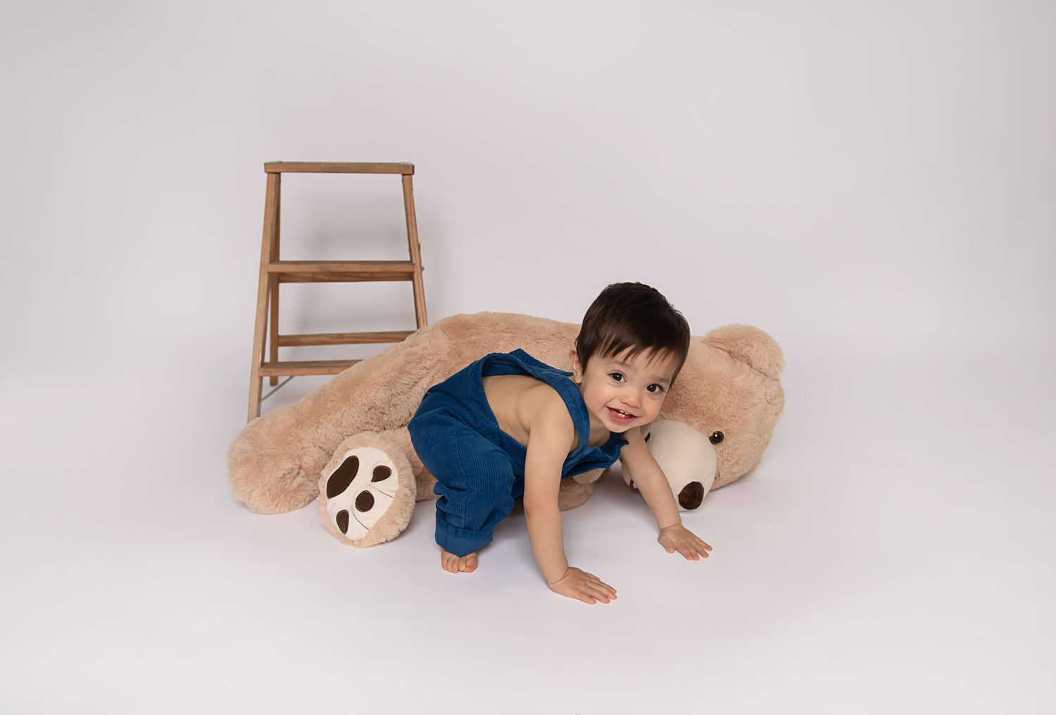 Small toddler in denim overalls playing with a large plush teddy bear on white background.