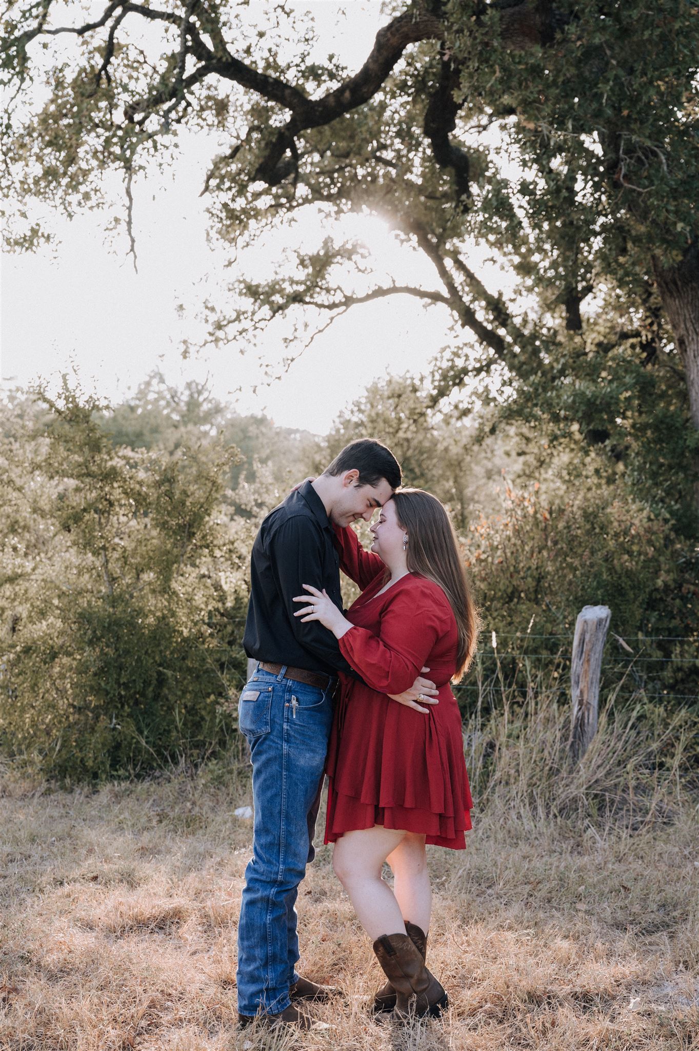 A romantic couple embraces under a sprawling oak tree during golden hour in a rustic outdoor setting.
