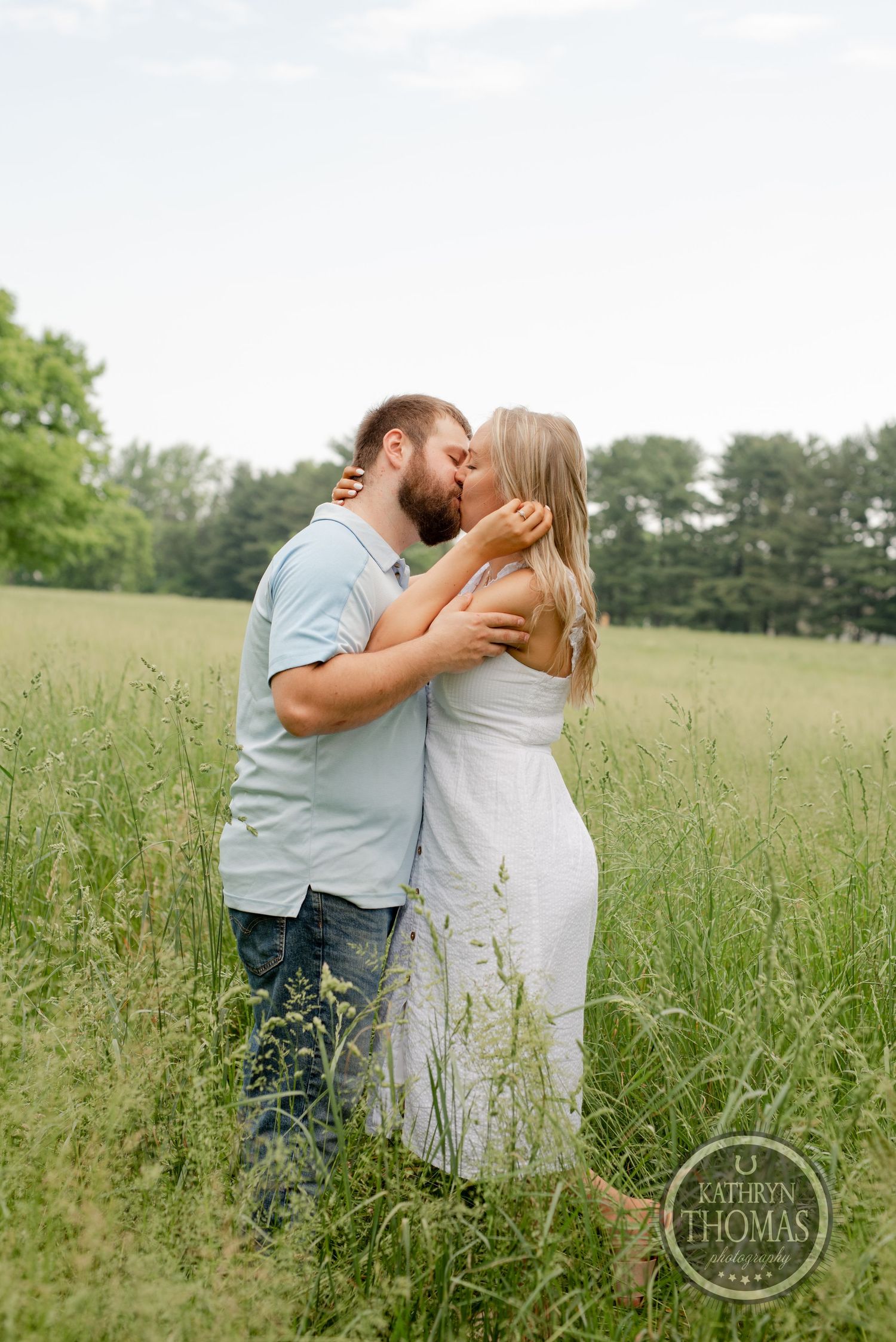 A passionate kiss shared between a couple standing in tall summer grass with blue sky above.