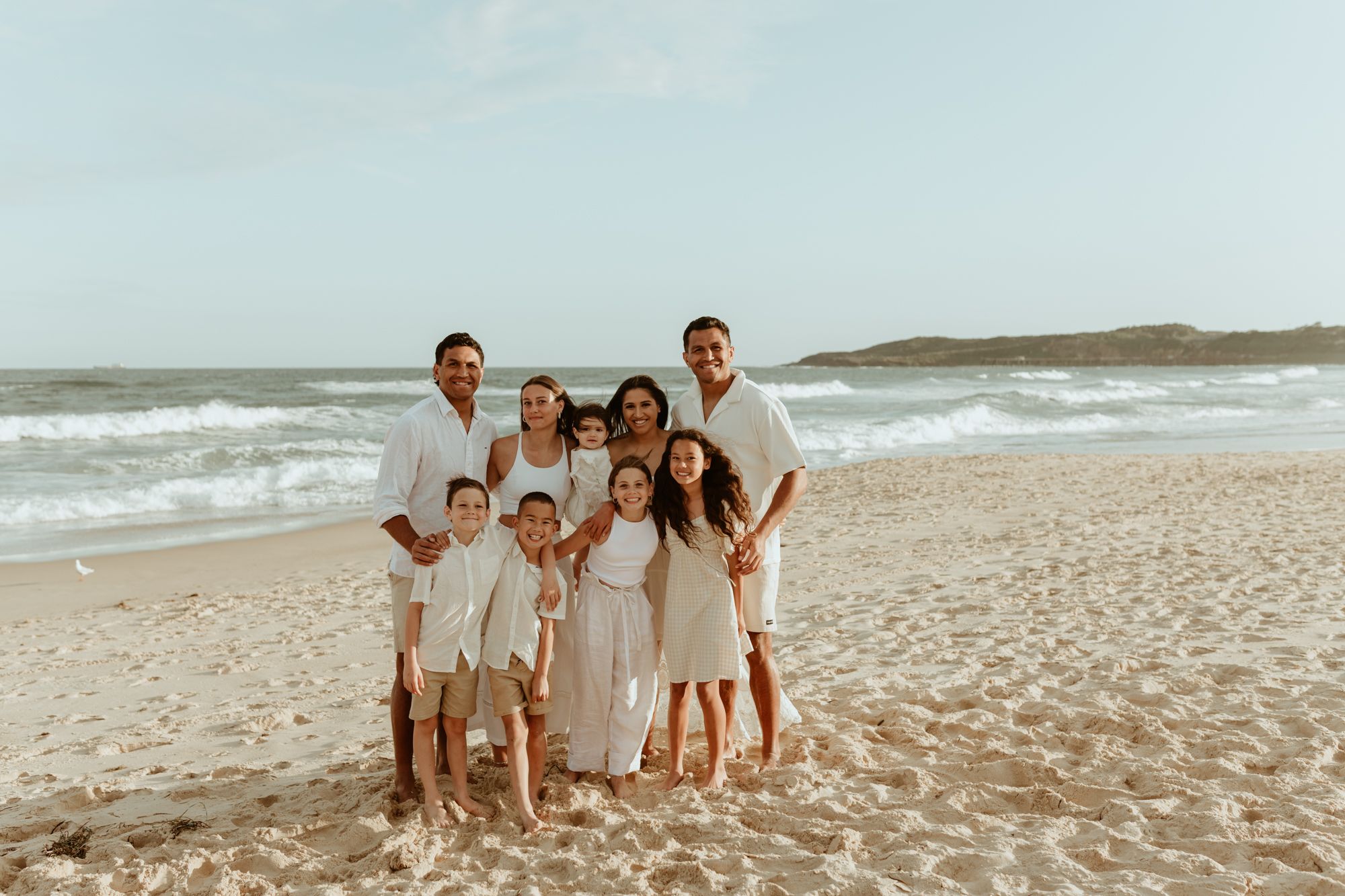 Family group in white and cream colored outfits poses together on sandy beach.