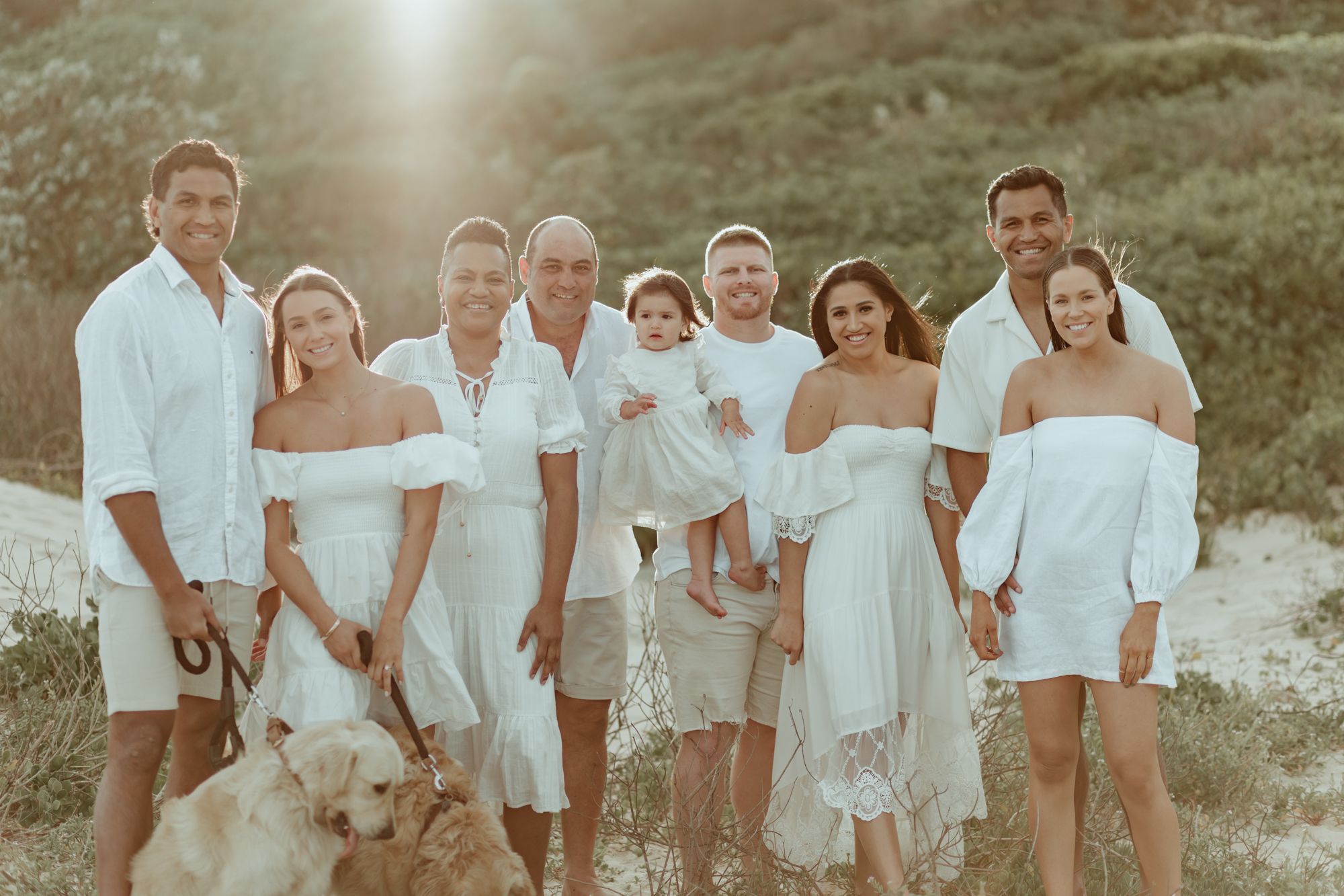 Family group in coordinated white outfits poses together at sunset on a coastal hillside.