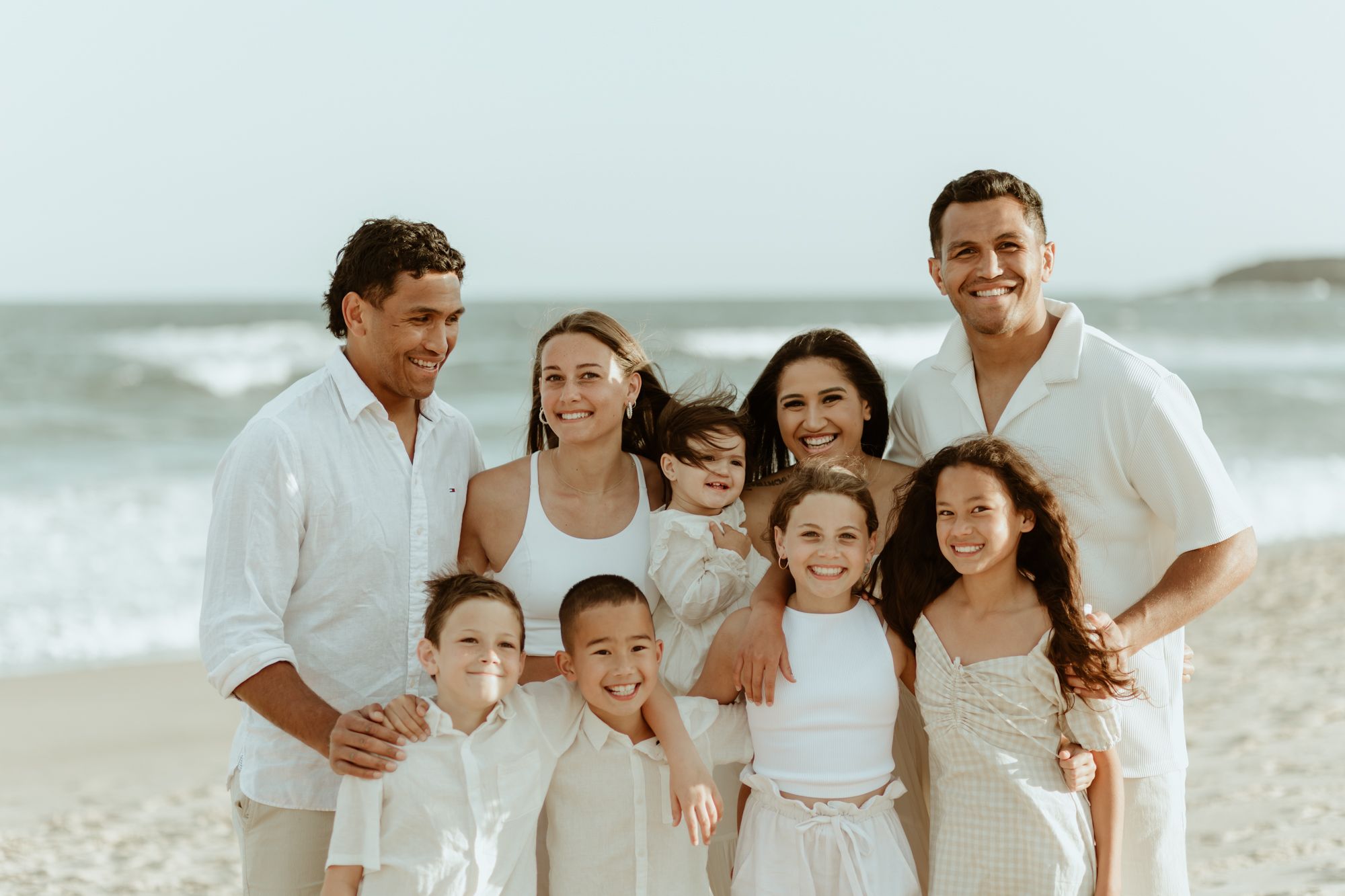 Multi-generational family wearing coordinated white outfits poses on beach with ocean waves behind them.