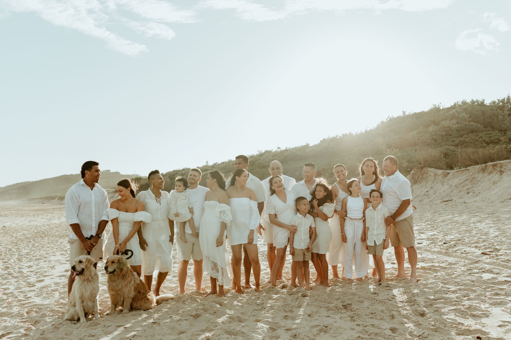 Extended family wearing white clothing poses together on beach with pets.