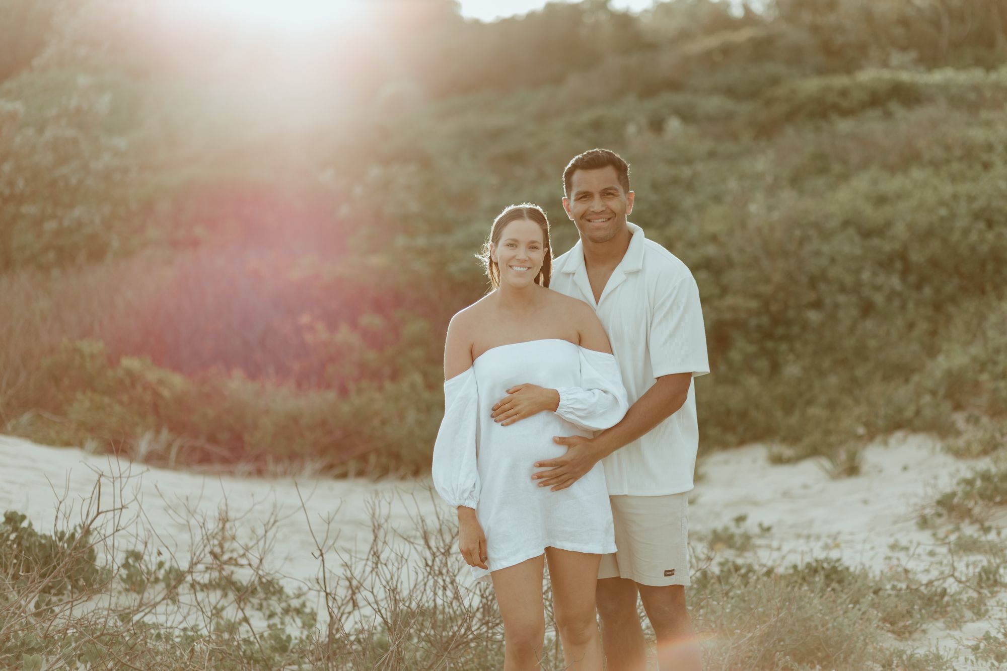 Couple in white clothing poses together during sunset on coastal hill.