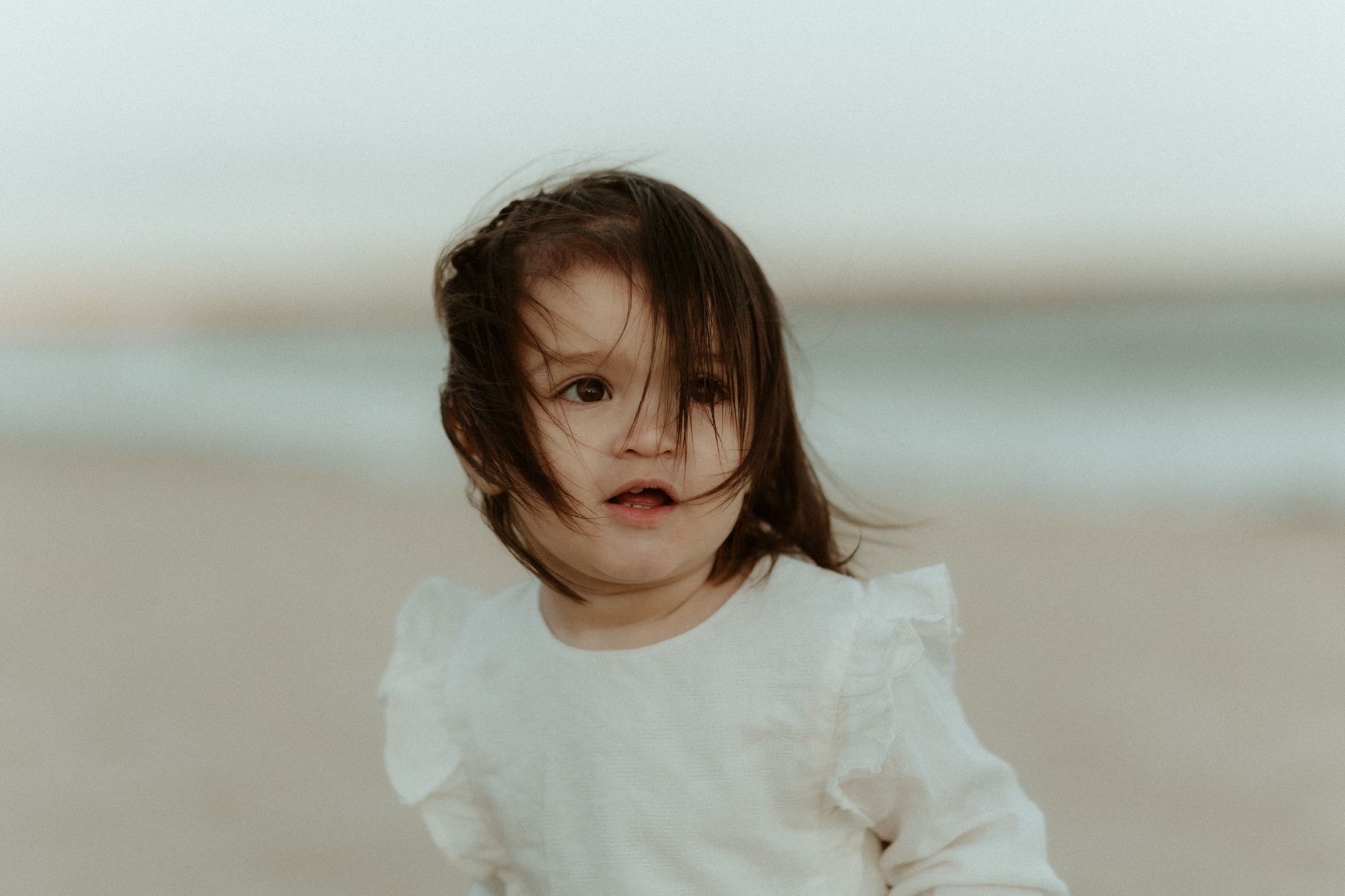 A small child in a white ruffled dress plays on a windy beach.