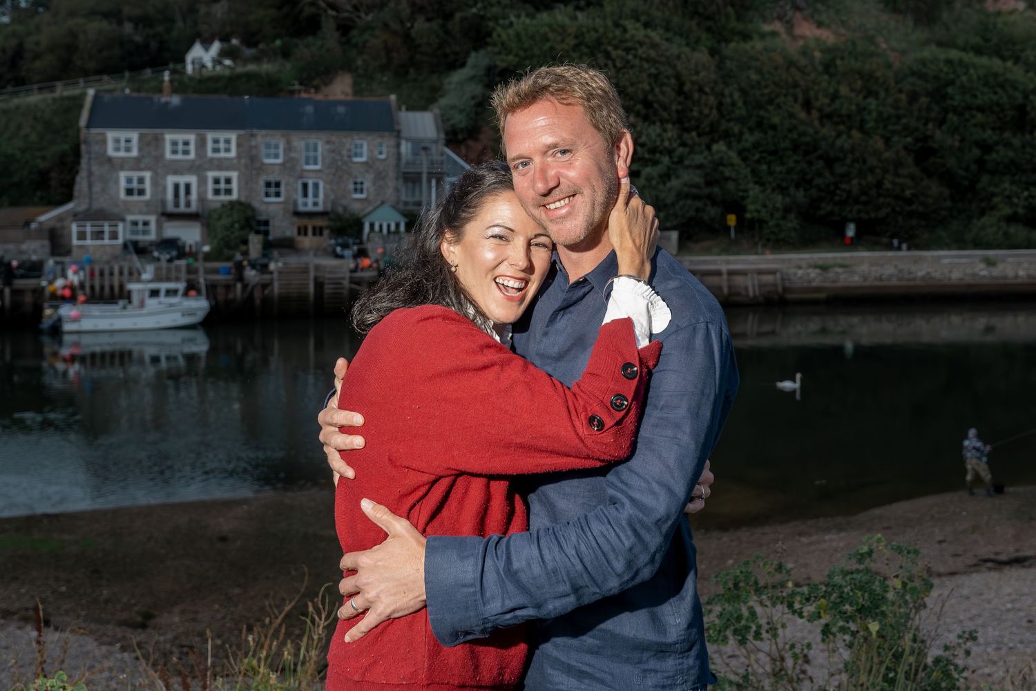 Couple embraces by waterfront at dusk with harbor houses in background.
