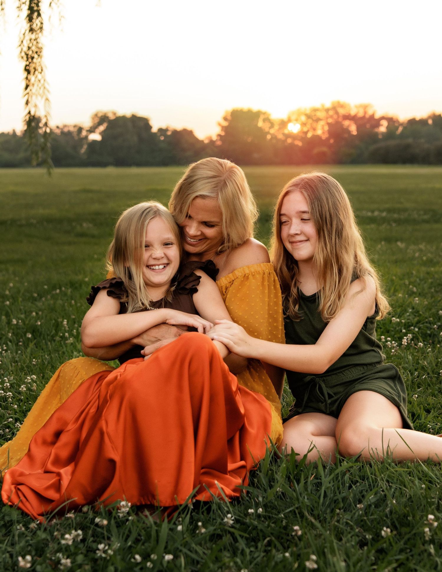 Mother in orange dress sits laughing with two daughters in green outfits on grass at golden hour sunset.