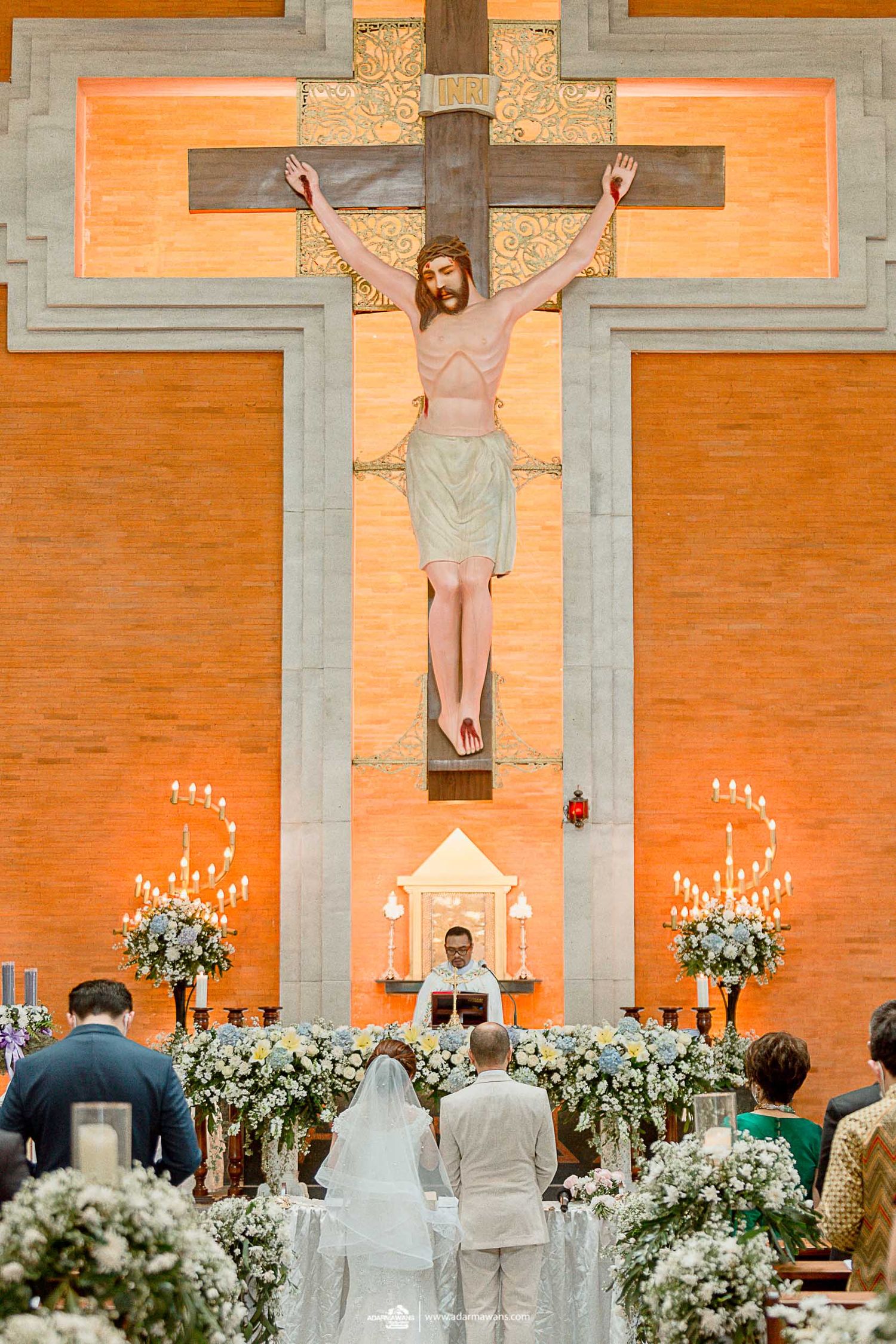 Wedding ceremony in front of large crucifix with white floral arrangements at Catholic church.