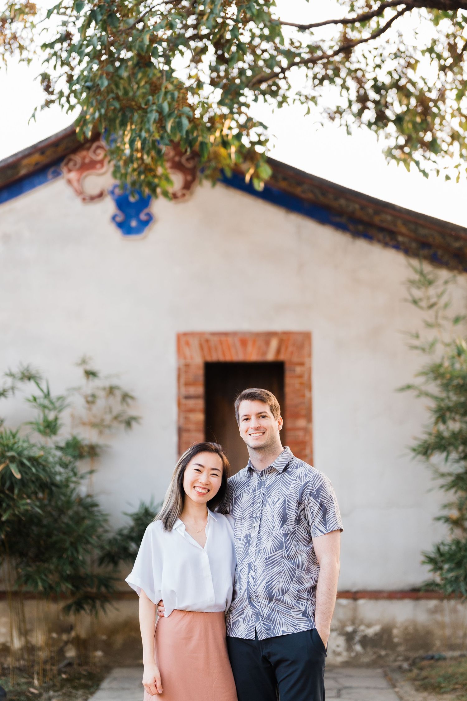 A couple poses in front of a white adobe building with rustic brick doorway and hanging vines.