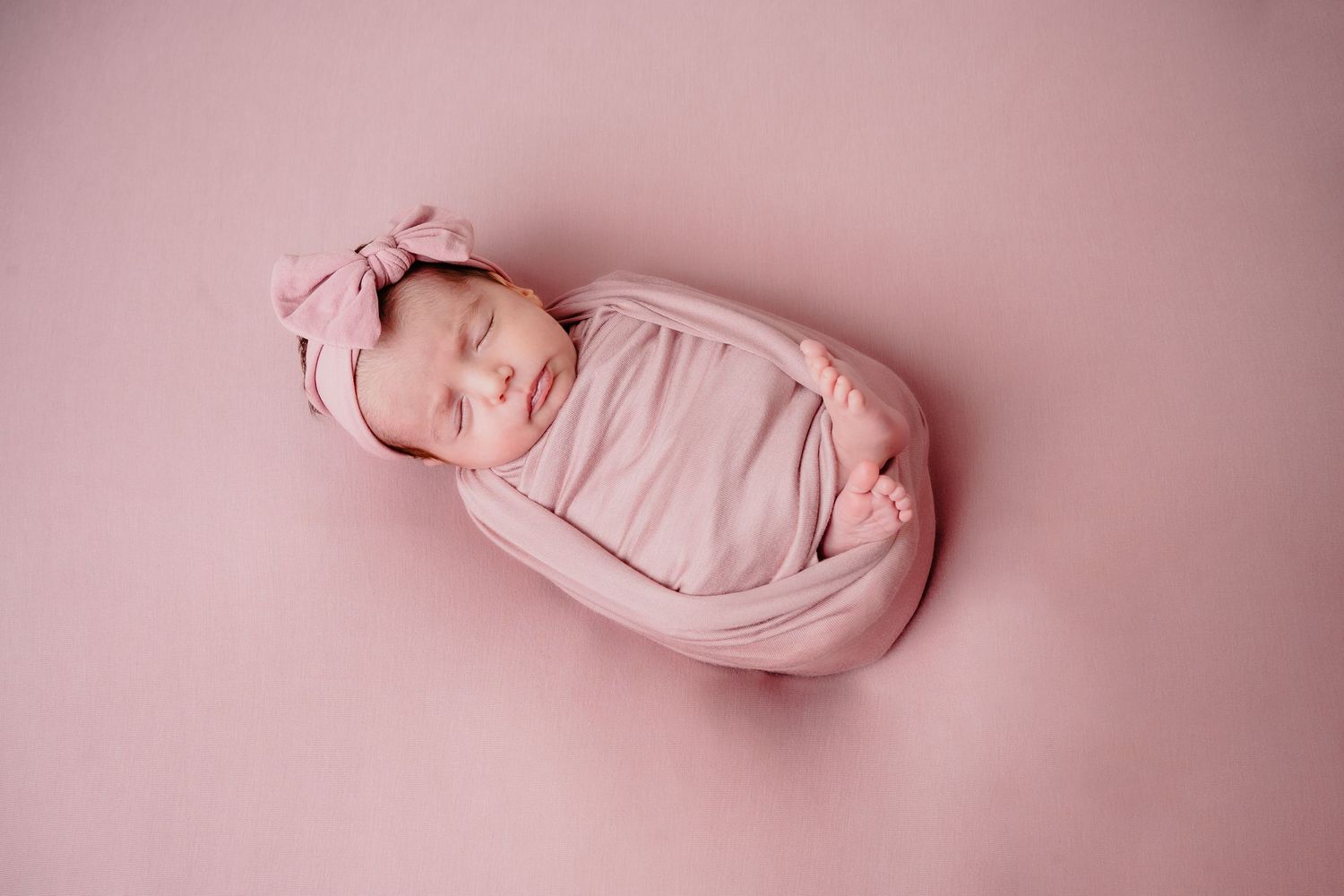 Sleeping newborn wrapped in pink swaddle with matching headband on pink background.