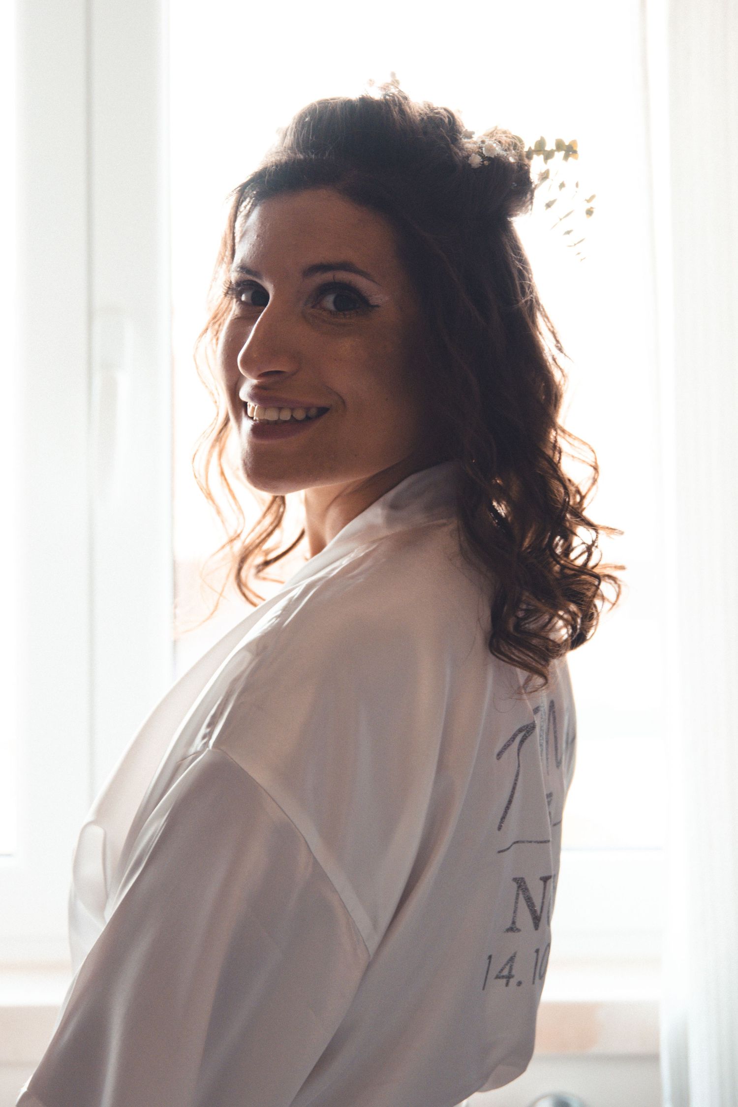 Bride in white robe smiles over shoulder during wedding day preparations.