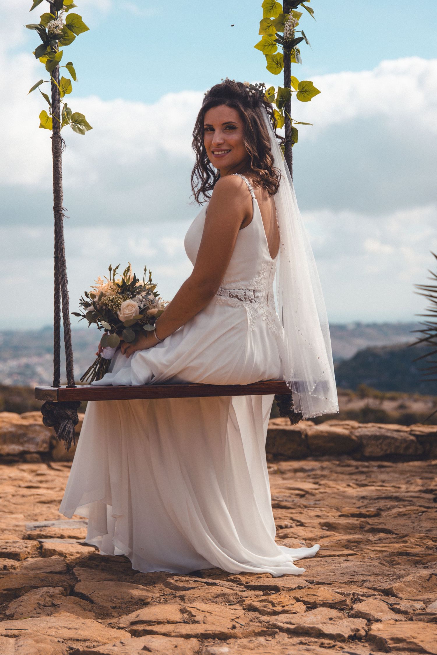 Bride in flowing white dress sits on rustic bench with mountain backdrop.