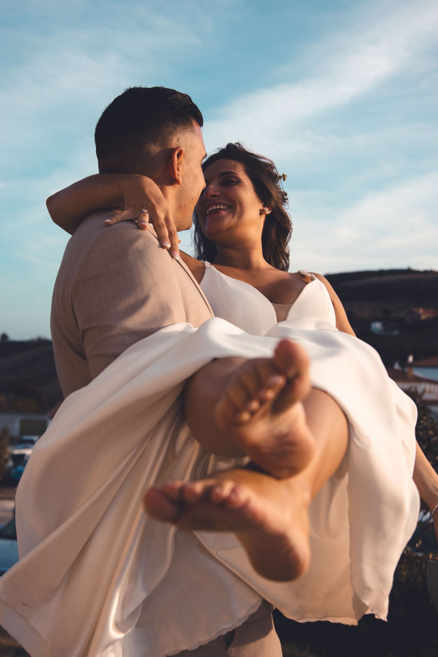 Groom in beige suit carries bride in white dress during sunset photo session.