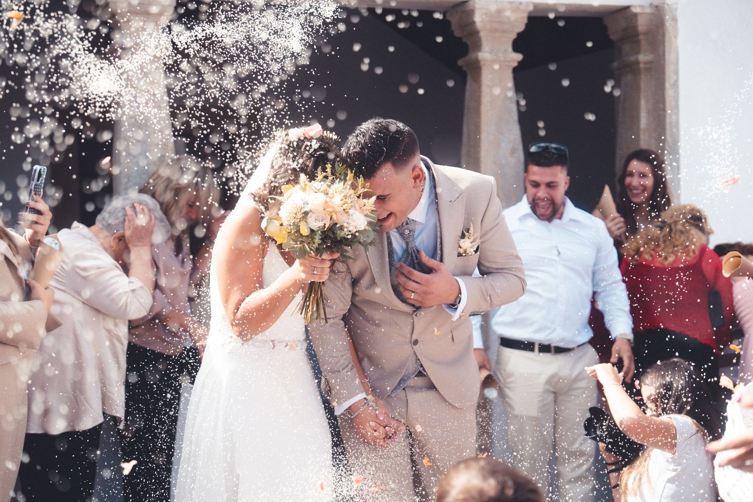 Newlyweds celebrate while guests throw confetti outside historic venue.
