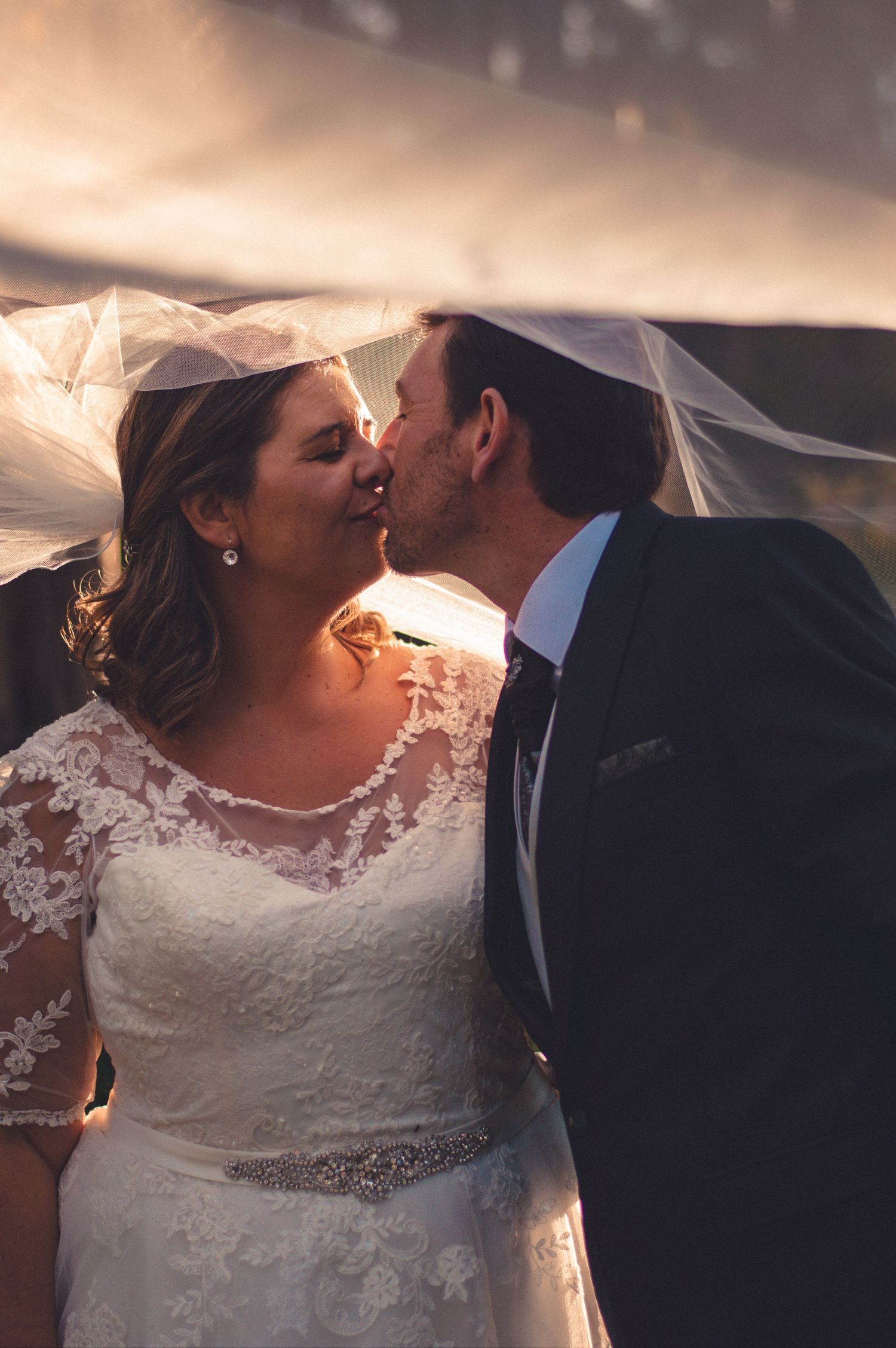 Romantic wedding moment captured under flowing veil at sunset.