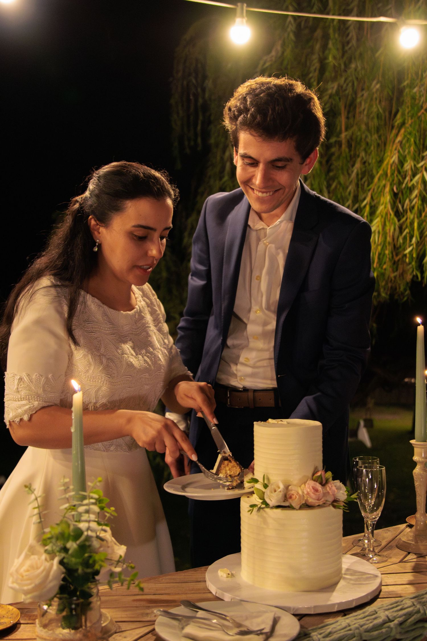 Newlyweds cutting their white wedding cake together at outdoor evening reception.