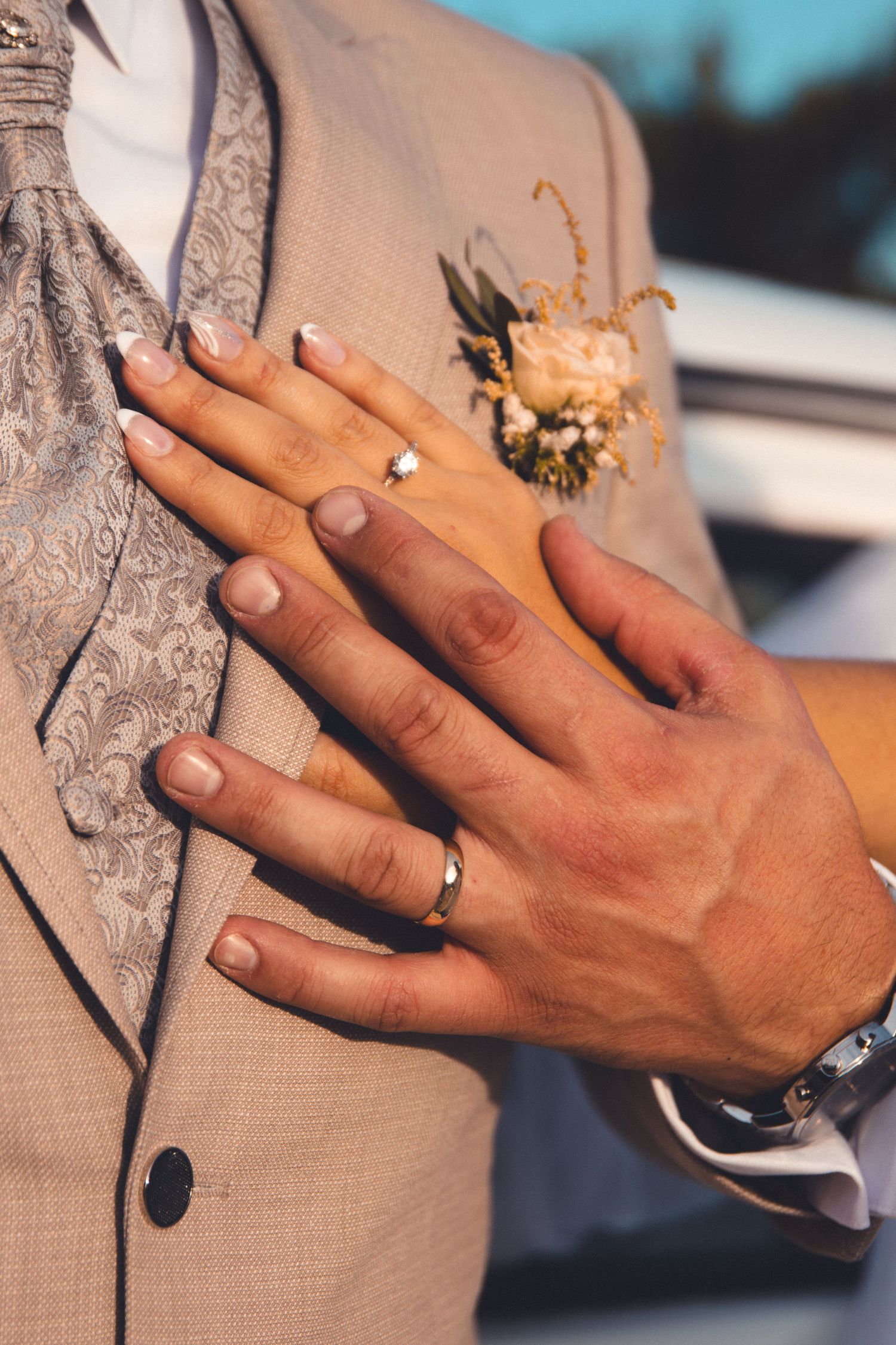 Close-up of wedding rings and boutonniere against beige suit jacket.