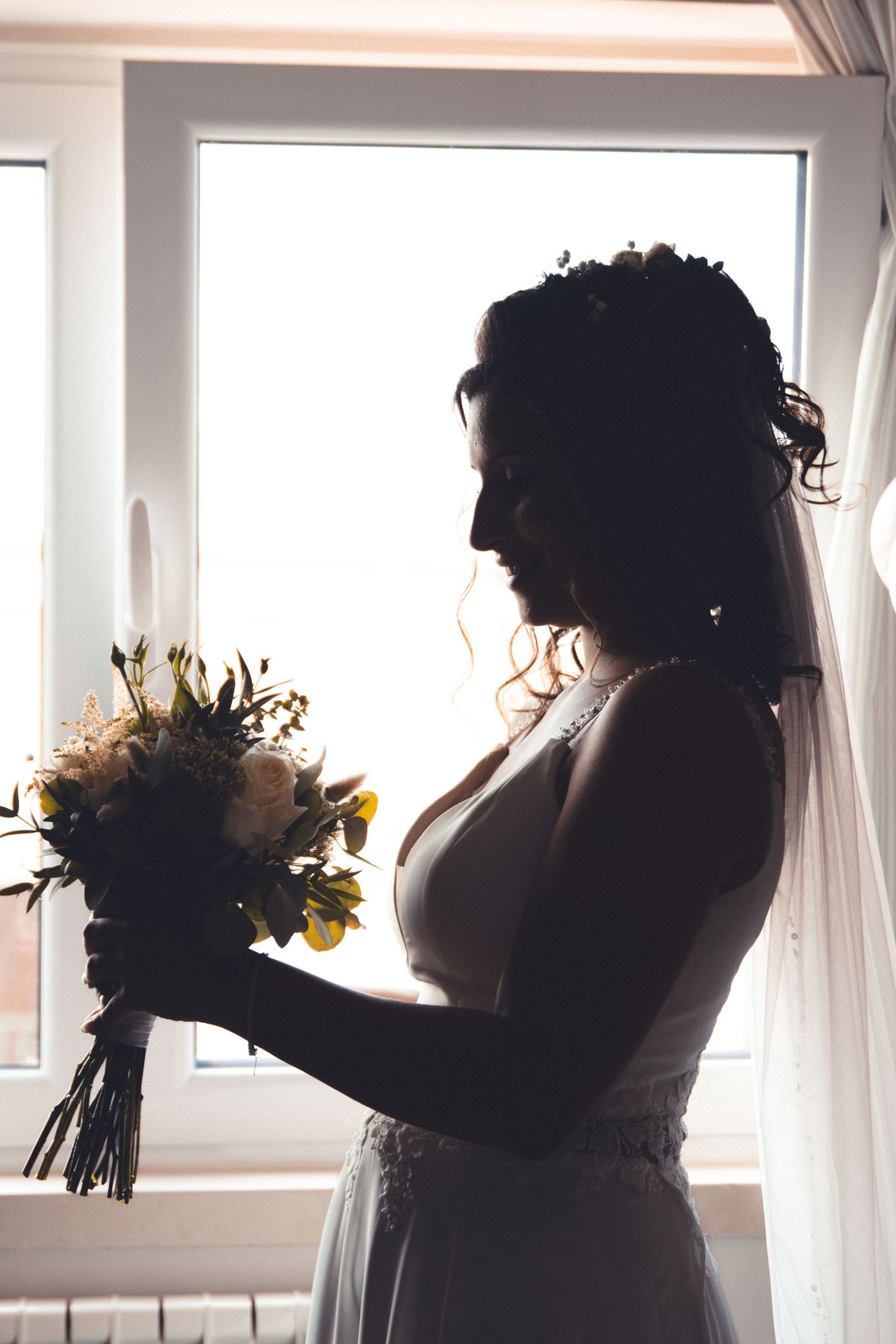 Silhouette of bride holding bouquet by window in dramatic lighting.