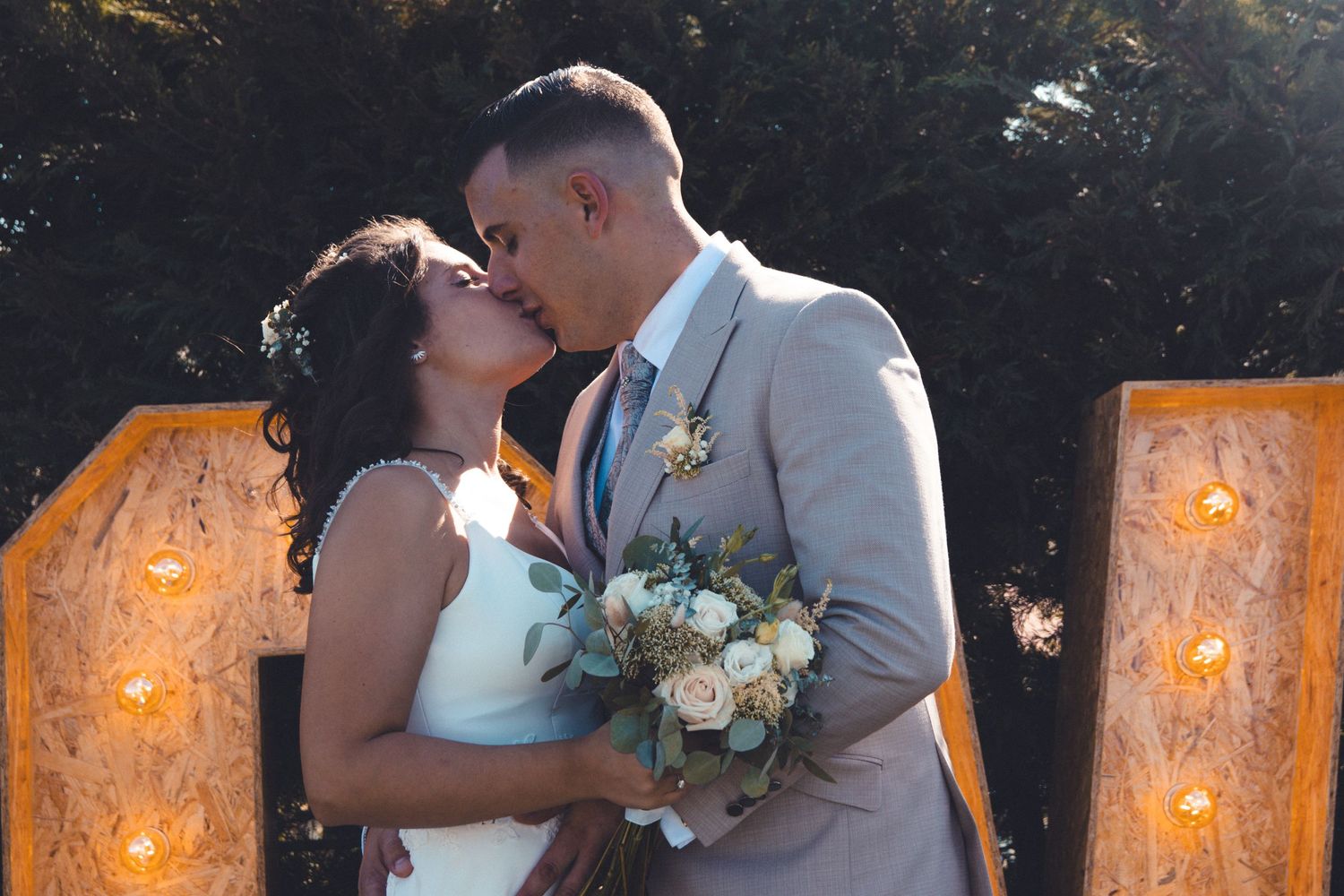 Newlyweds share romantic kiss in front of illuminated rustic backdrop.