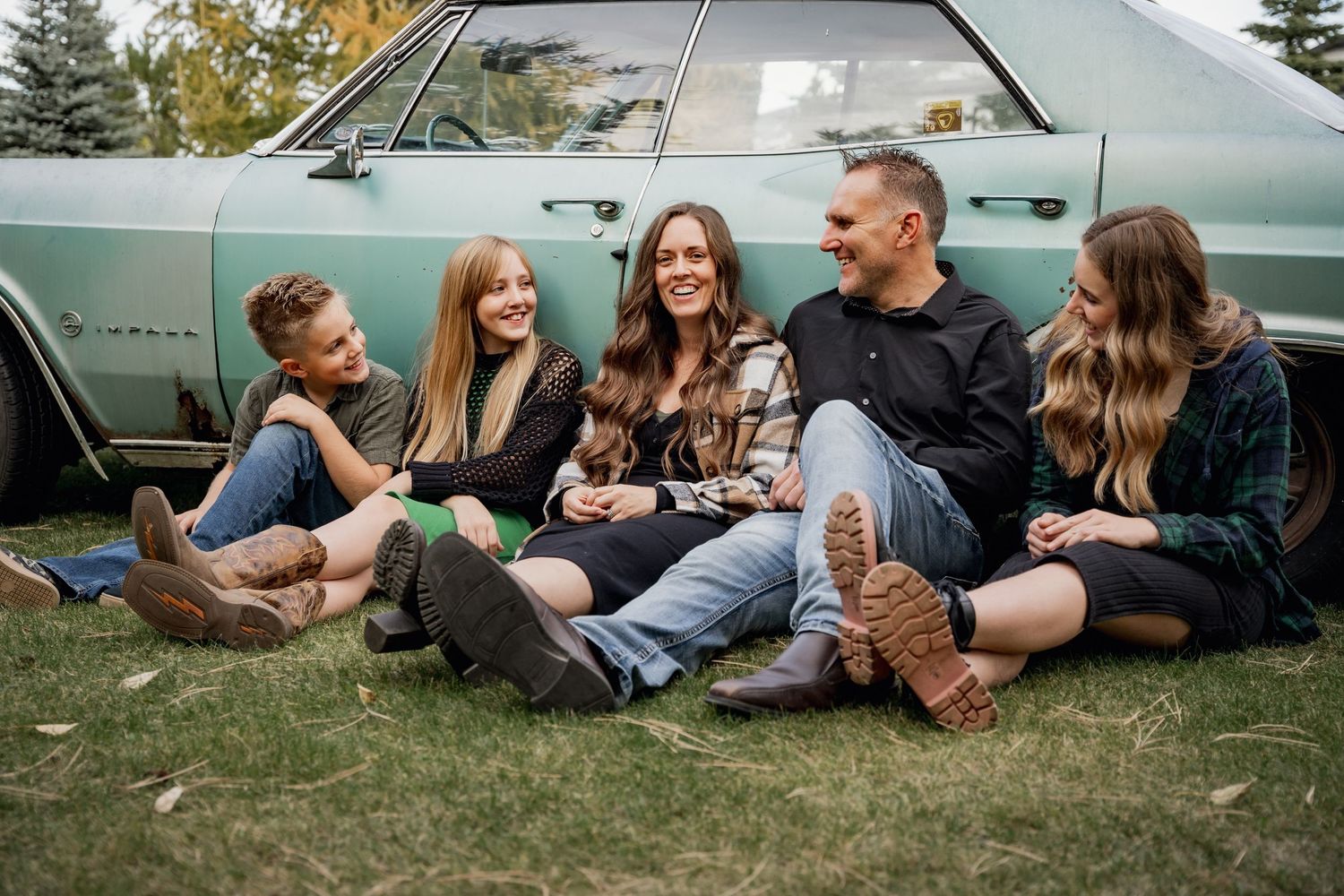 Group relaxing on grass beside vintage turquoise car during casual outdoor photoshoot.