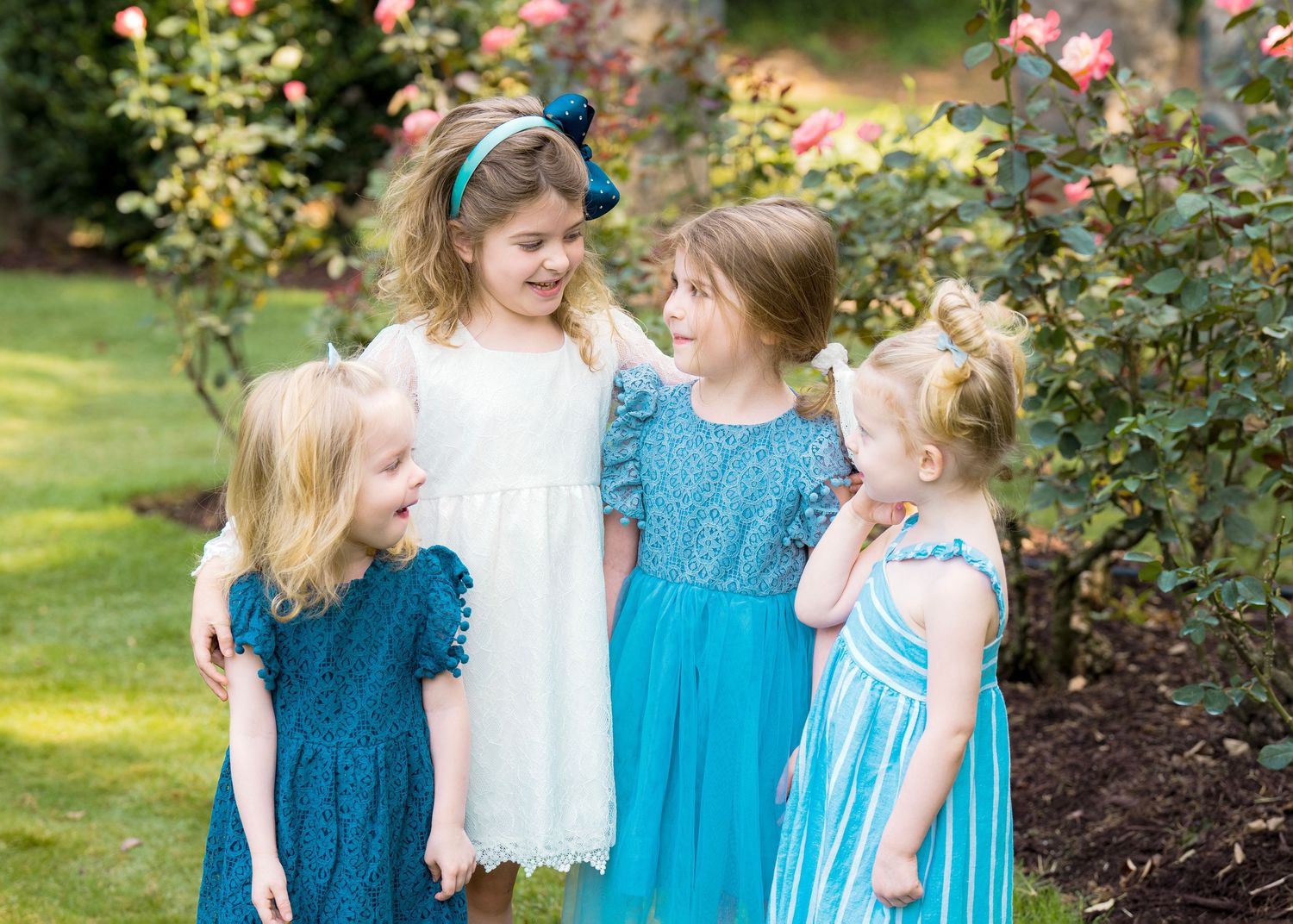 Four young girls in blue and white dresses smile together near blooming rose bushes in a garden.