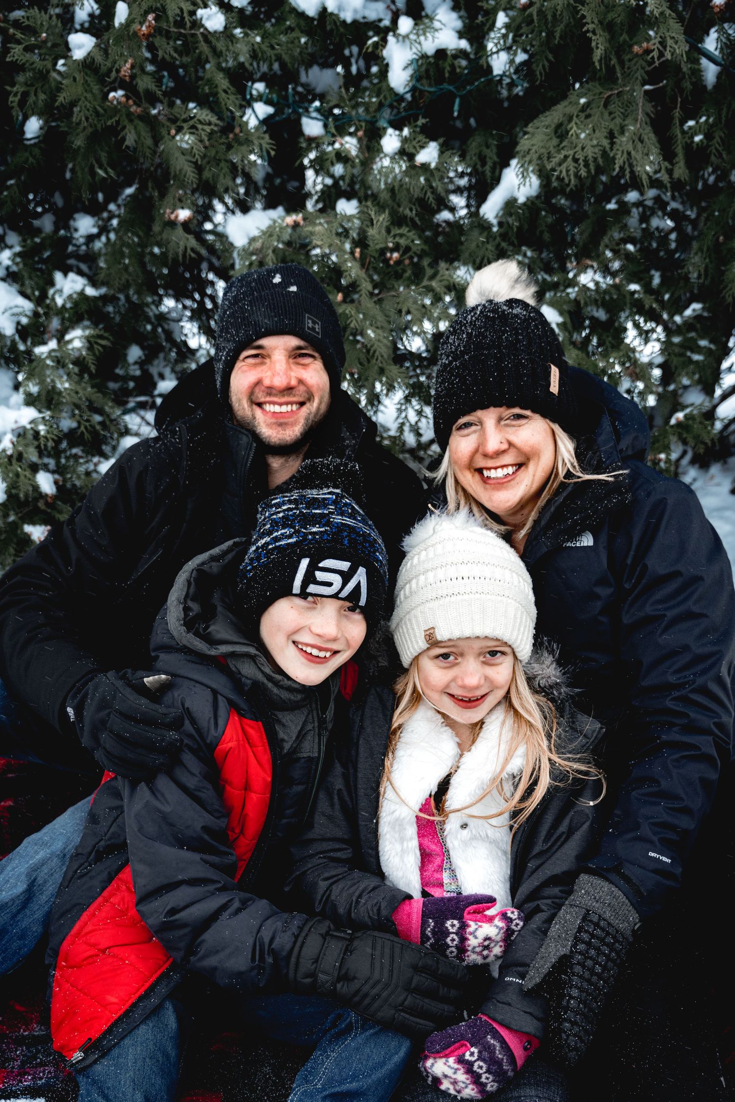 Happy family wearing winter clothes and knit hats posing together in snowy evergreen setting.