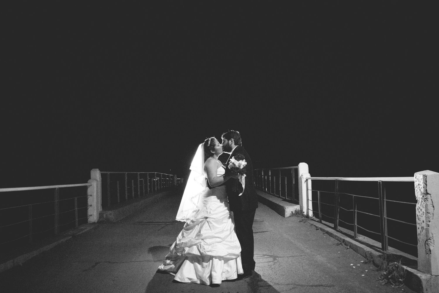 Romantic black and white photo of newlyweds embracing on a bridge at night.