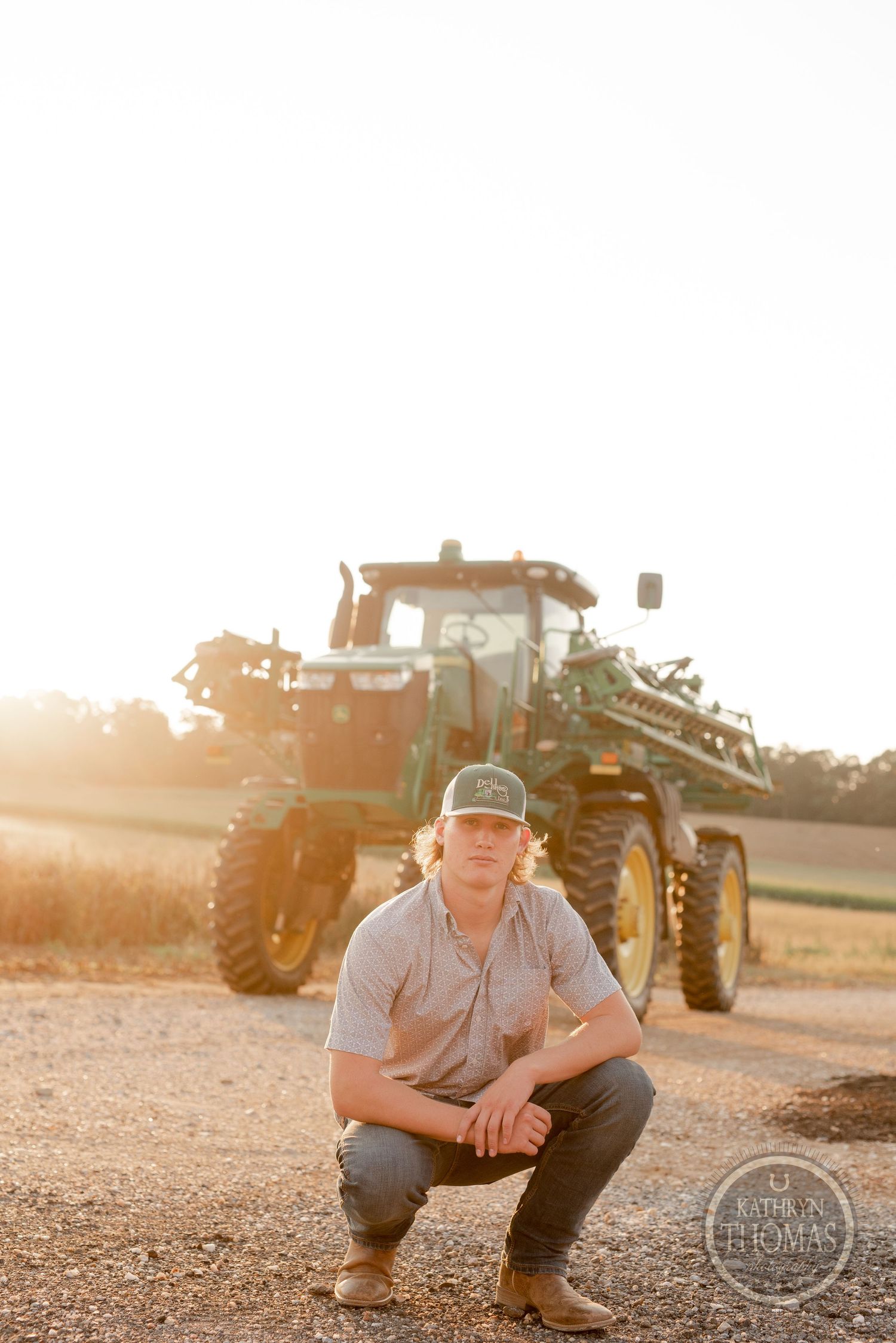 Farmer crouches in front of a John Deere tractor during sunset on a dirt path.
