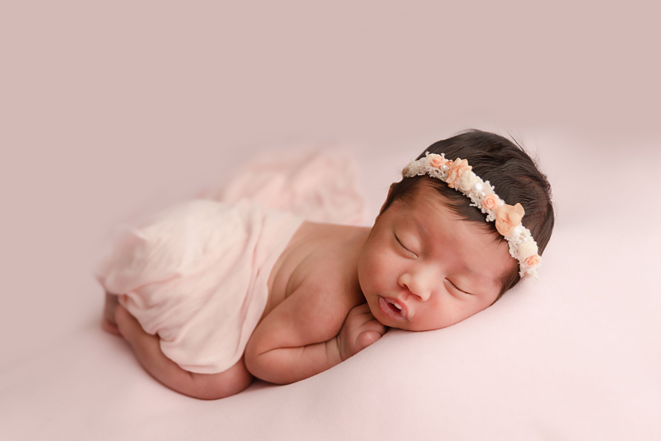 Newborn baby girl sleeping peacefully wrapped in pink fabric with a floral headband on a soft pink background.