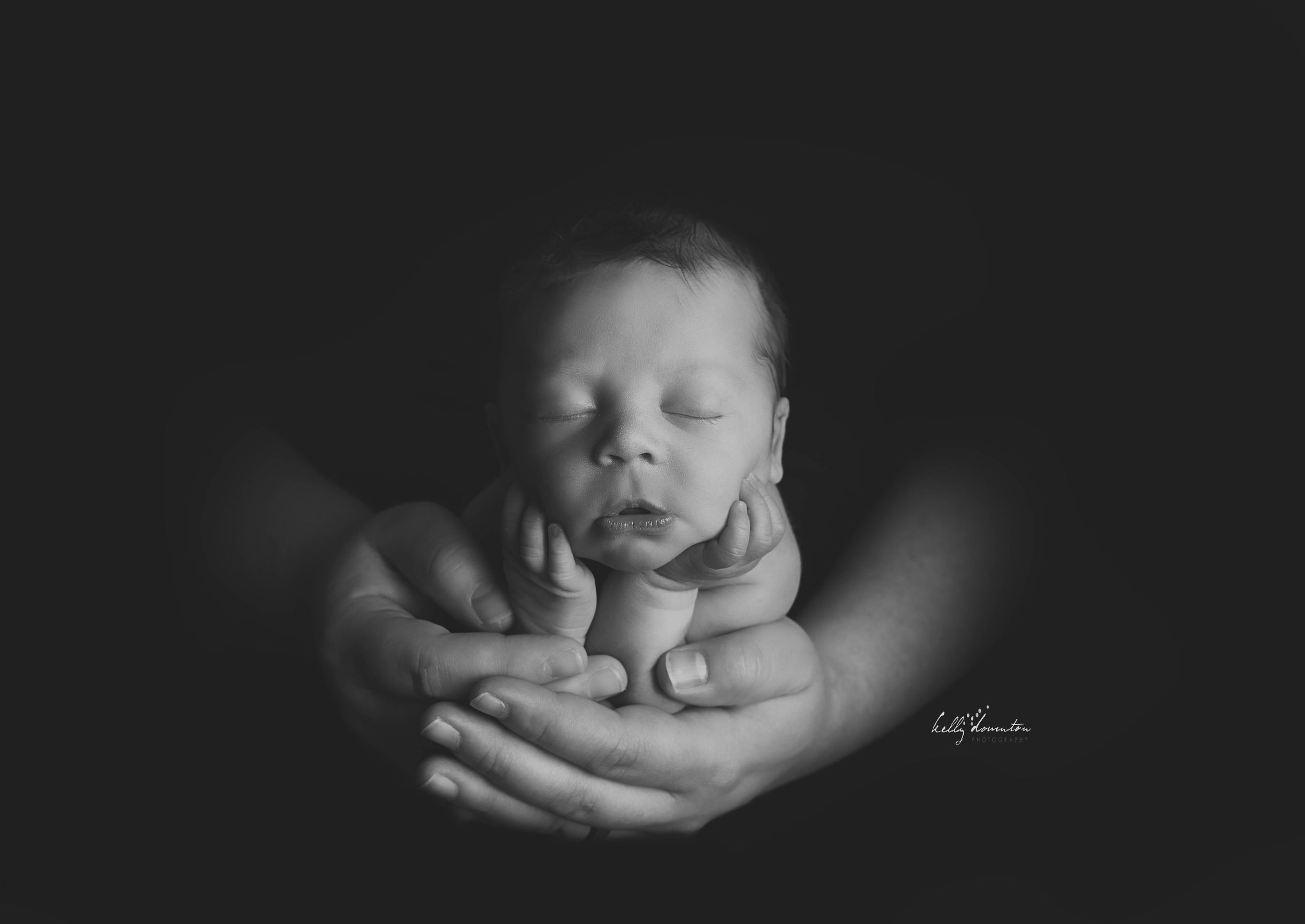 Black and white newborn photography of sleeping baby cradled in adult hands against dark background.