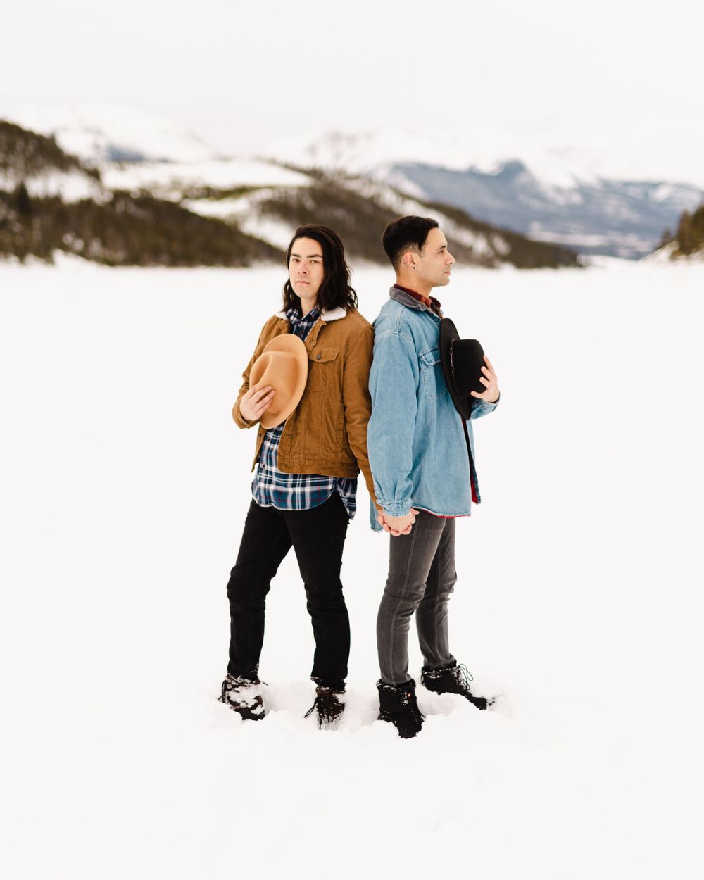 Two people in winter jackets stand back to back on a snowy mountainside landscape.
