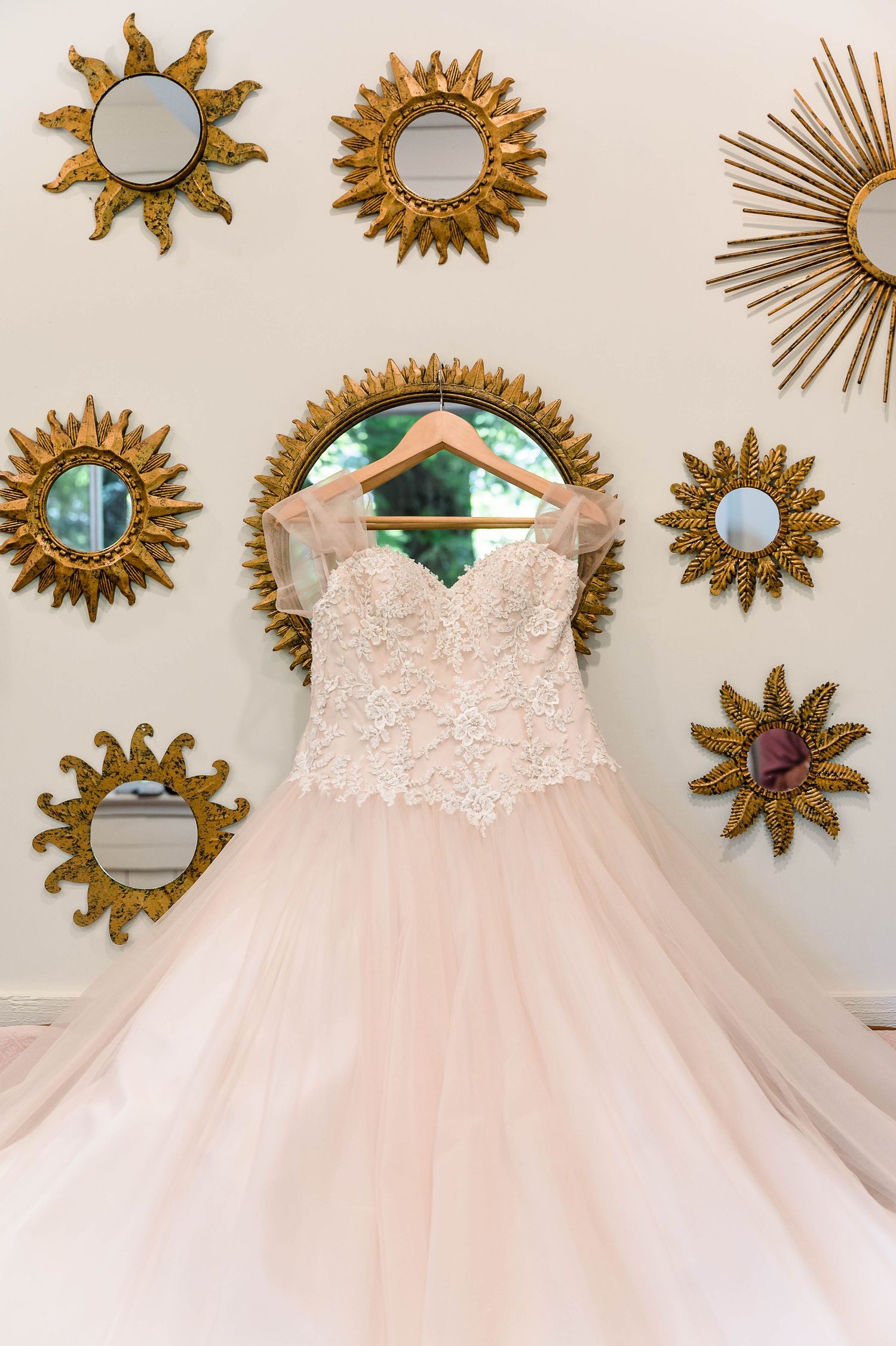 Ethereal blush wedding gown hanging among golden sunburst mirrors in preparation room.