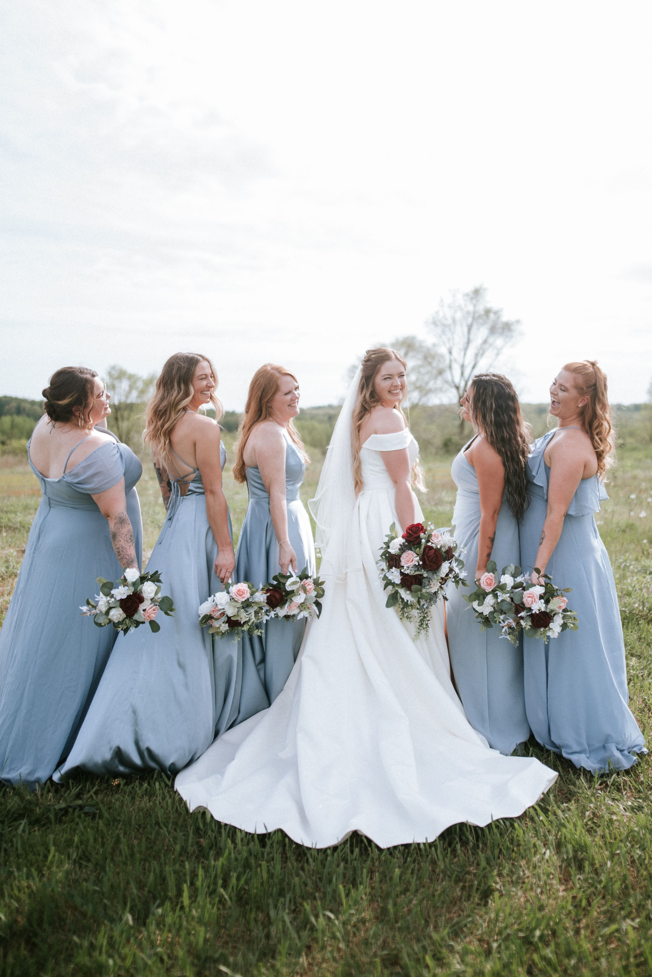 Bride and bridesmaids in powder blue dresses show off bouquets in candid outdoor photo.